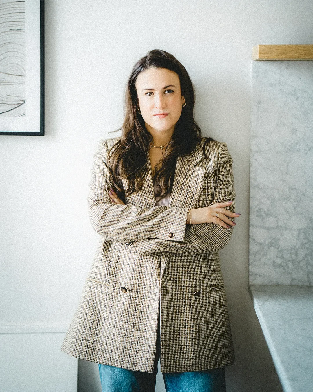 A photo of Carly Jenner, a woman with long dark hair stands with her arms crossed, wearing a beige plaid blazer and blue jeans in a modern, well-lit room with minimalist decor.