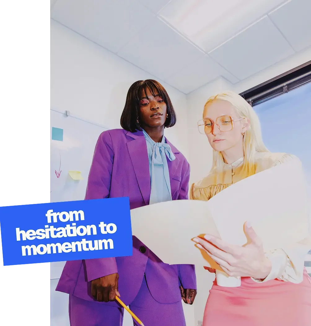 Two women, one with dark skin wearing a purple suit and the other with light skin and blonde hair wearing glasses and a light-colored blouse, look at a large sheet of paper together inside an office with a window showing a blue sky.