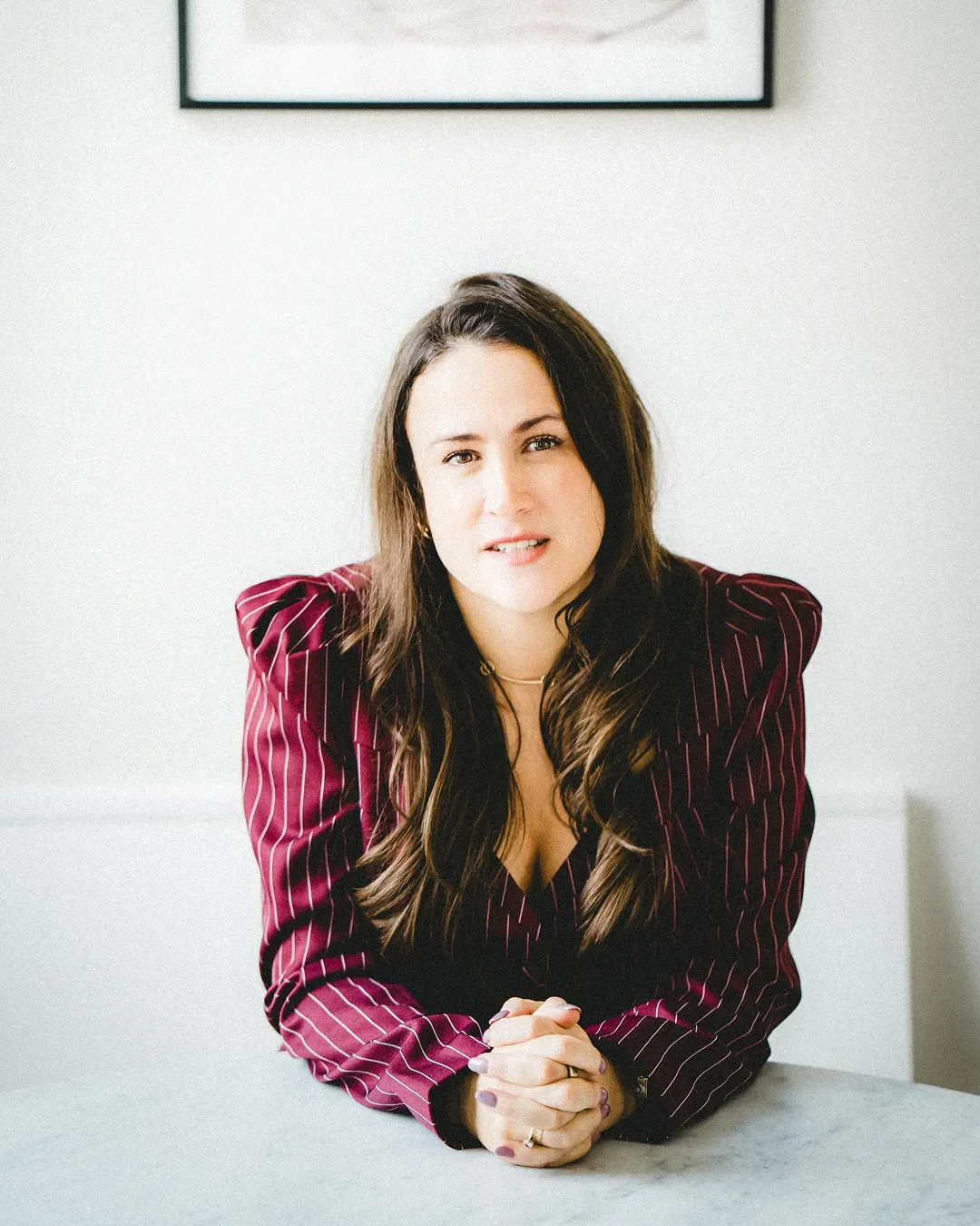 A woman with long brown hair, wearing a maroon pinstripe blazer, is sitting at a table with her hands clasped, looking at the camera with a slight smile. There is a framed picture on the white wall behind her.
