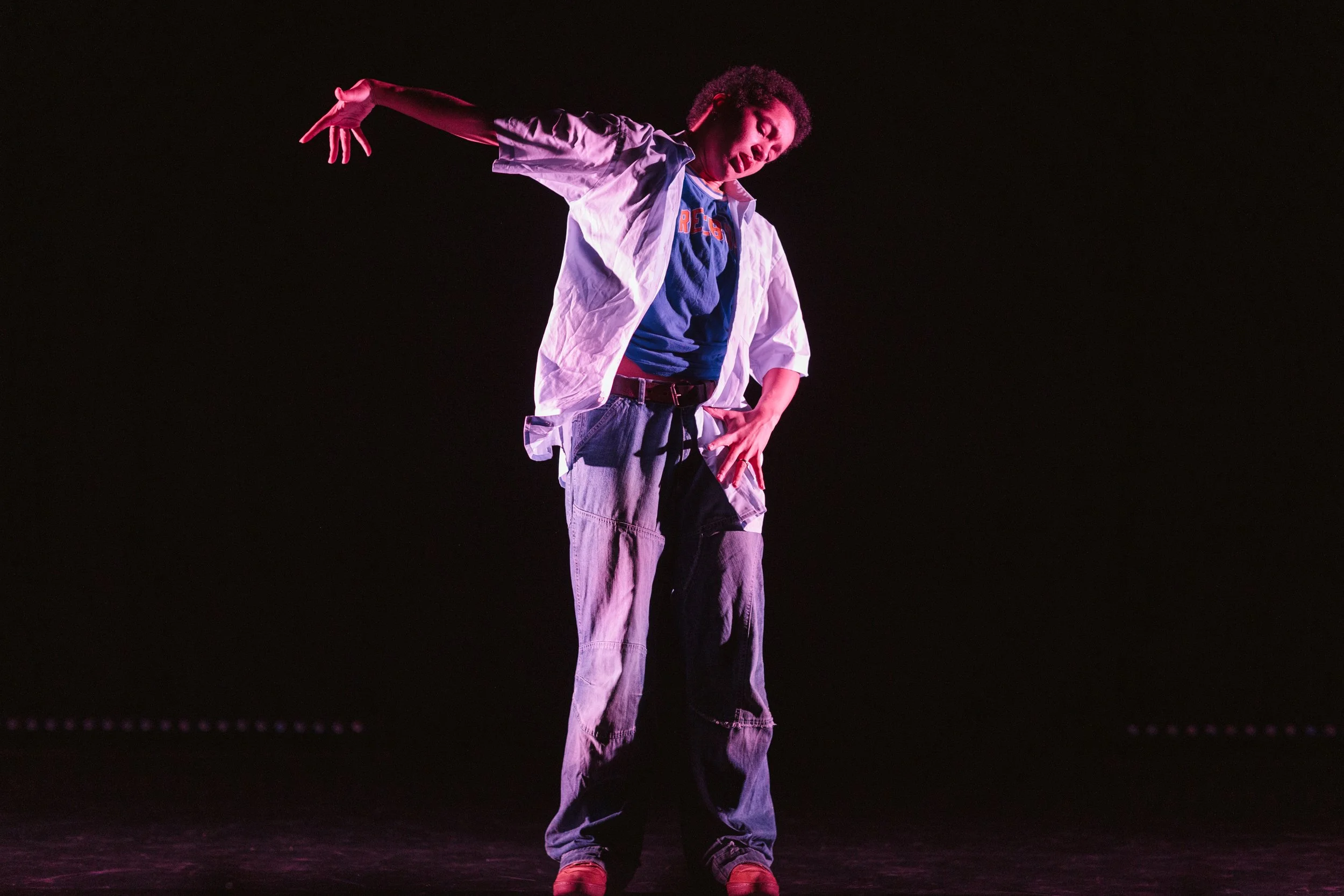 A person performing dance on stage, wearing a white open shirt, blue t-shirt, baggy jeans, and red shoes, with one arm extended and hand near the face, against a dark background.
