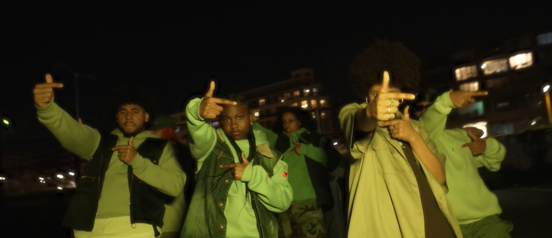 Group of young people at night, wearing green and beige clothing, pointing finger guns at the camera, with city buildings in the background.