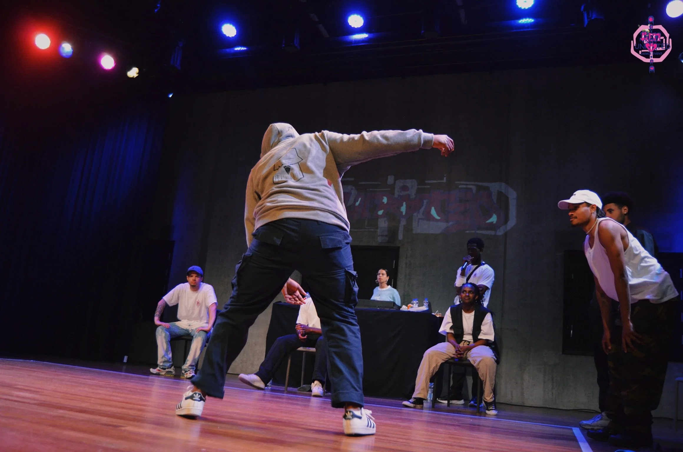People participating in a dance battle on stage, with a person in streetwear performing, and others watching or judging in the background.