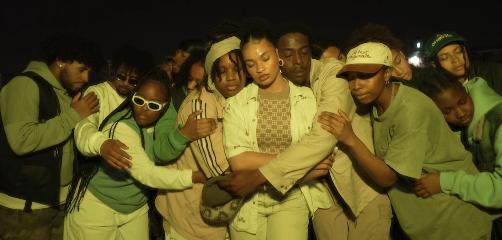 A group of diverse young people gathered in a circle, with their eyes closed and arms around each other, participating in a nighttime group prayer or meditation outside.