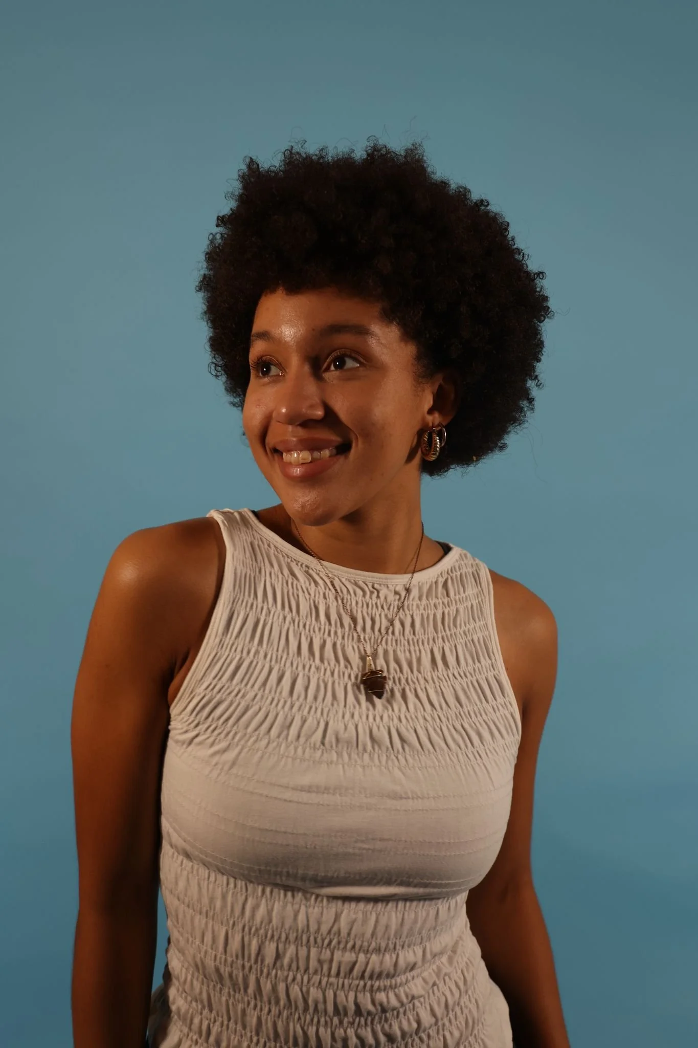 A woman with a large afro hairstyle smiling, wearing gold earrings, a necklace with a pendant, and a sleeveless beige top with textured fabric, against a solid blue background.