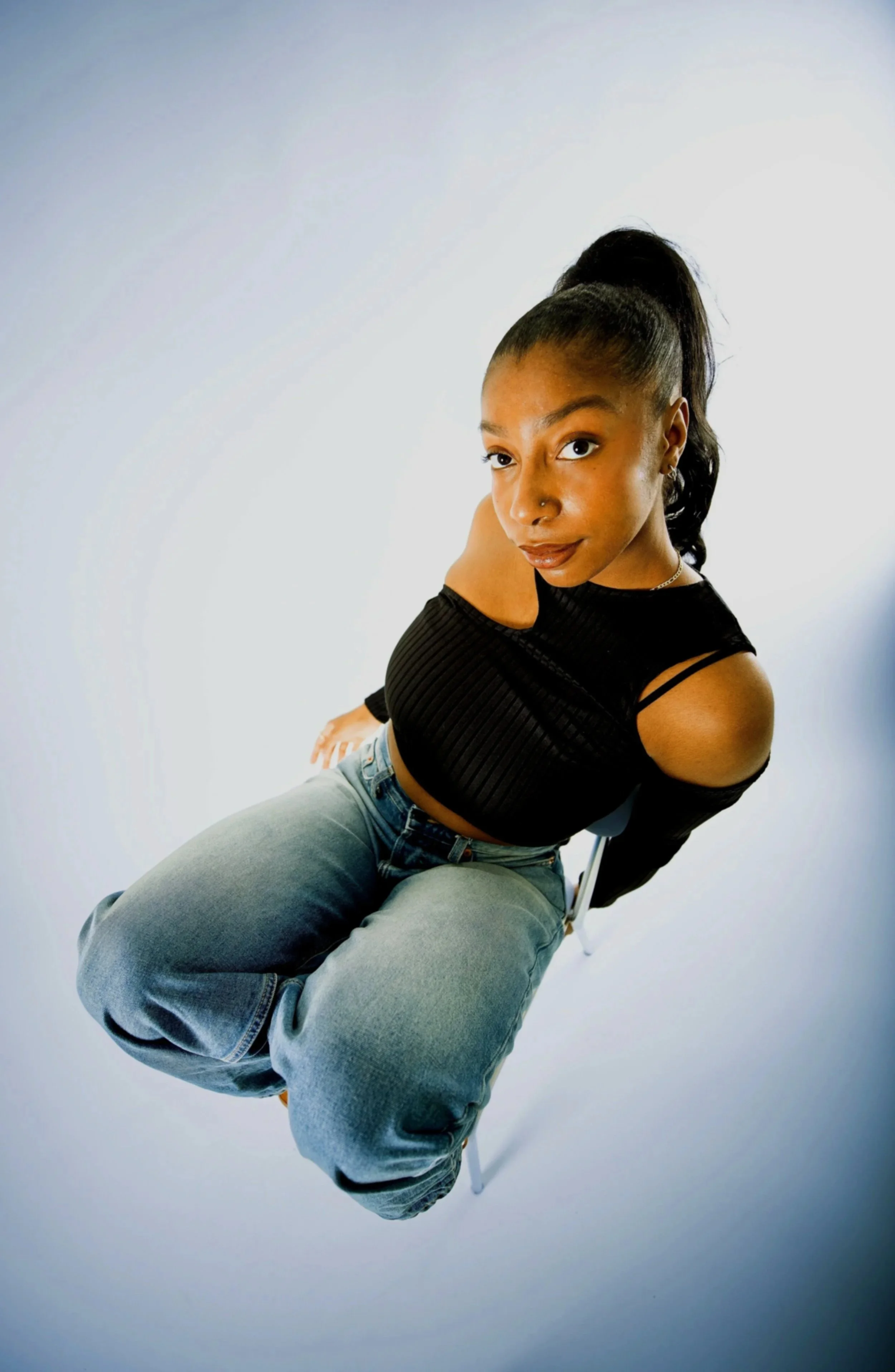 A young woman with dark hair in a high ponytail is sitting on a chair, wearing a black cutout top and light blue jeans, looking directly at the camera.
