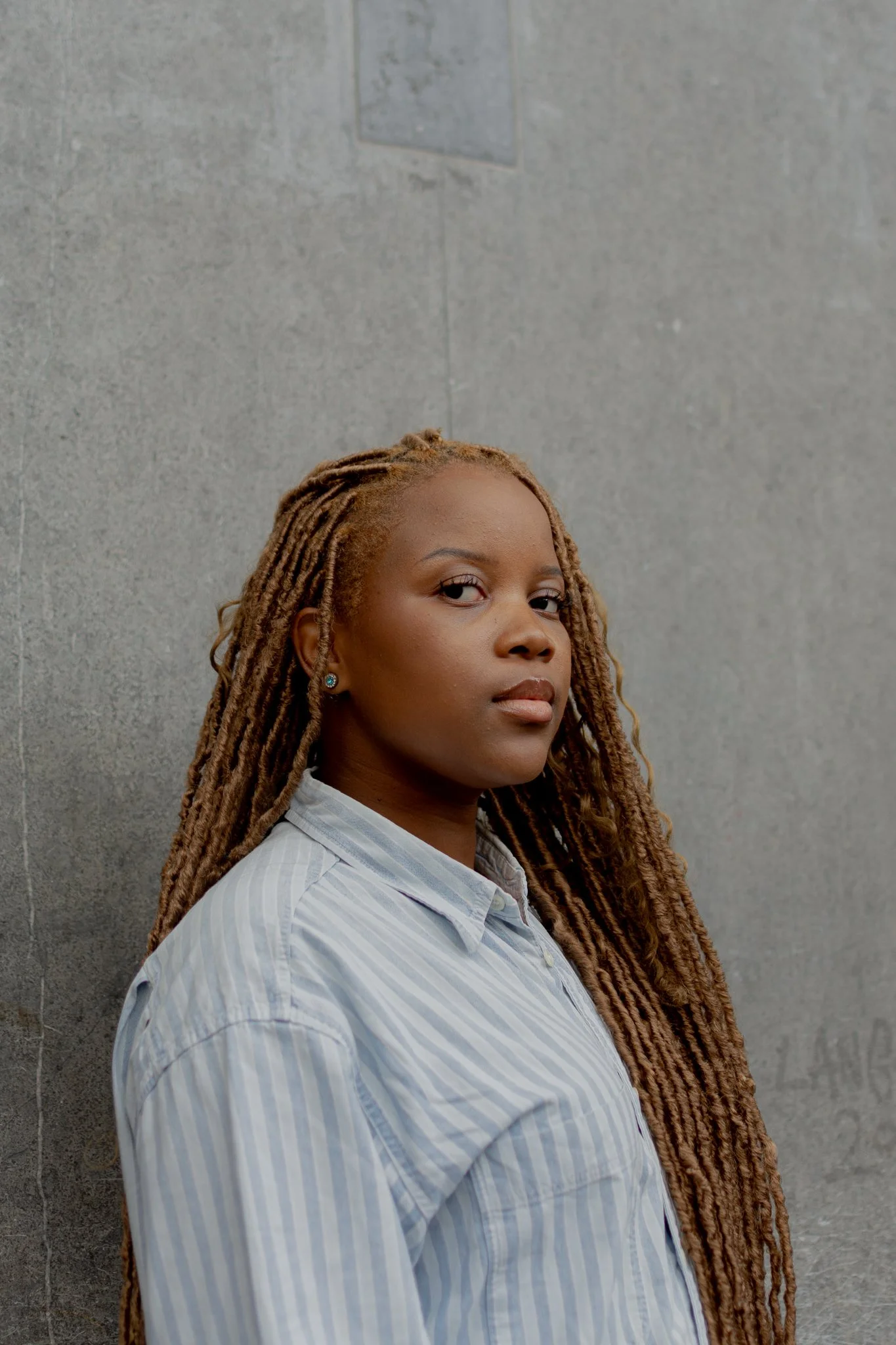A young woman with long, light brown dreadlocks, wearing a light blue and white striped shirt, standing against a gray concrete wall, looking at the camera.