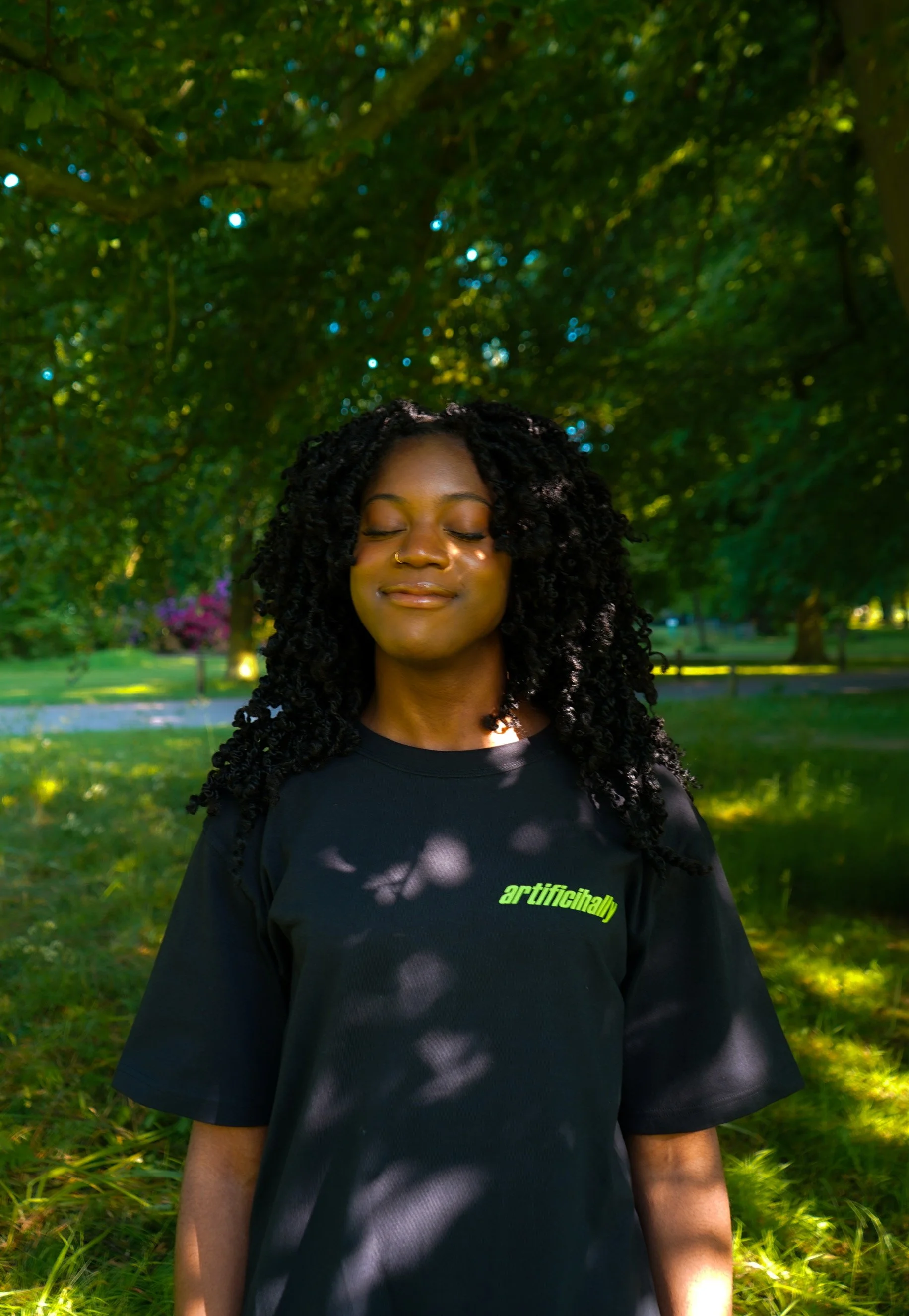 A woman with curly black hair and closed eyes standing outdoors in a park with green trees and grass, wearing a black t-shirt with the word 'artificihally' in green text.