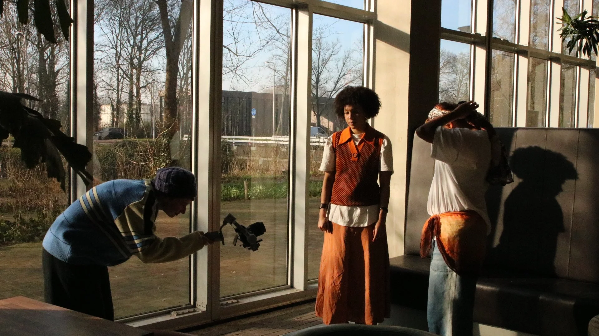 A woman is filming with a camera, two women are sitting near the glass windows, one standing in front of the window, all inside a building with large glass windows showing trees and outdoor scenery.