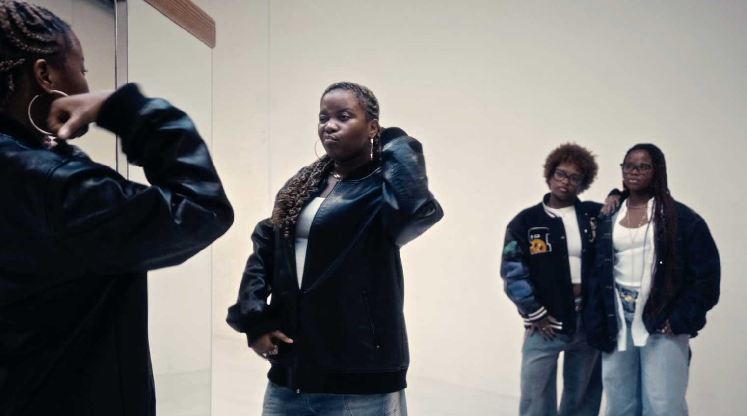 Four women standing in front of a mirror in a plain room, two of them posing with one arm on their side and another with an arm over a friend's shoulder, all dressed casually in jackets and jeans.