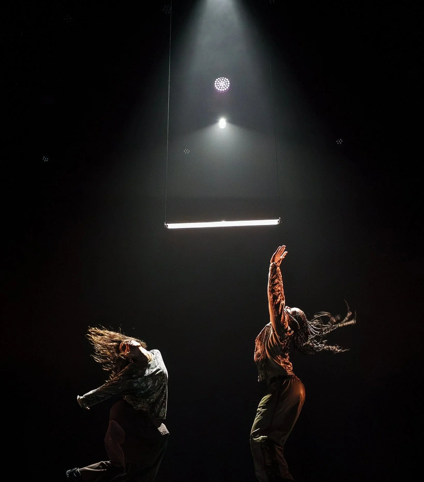 Two dancers on stage illuminated by a spotlight, with a dark background and minimal lighting fixtures above.