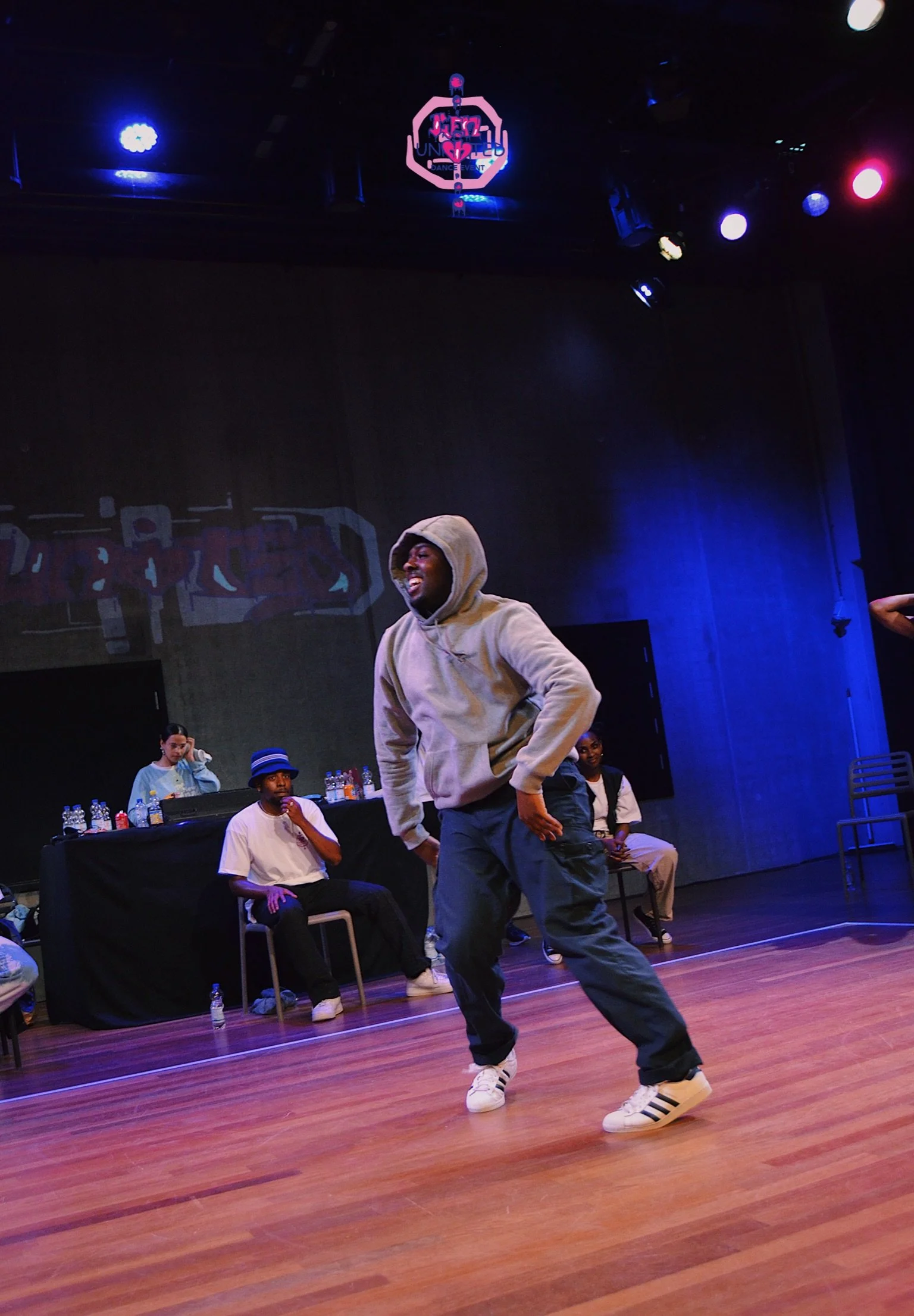 A man dancing on stage with a gray hoodie and black pants, surrounded by others sitting behind him, with a DJ and water bottles on a table in the background, colorful stage lights overhead.