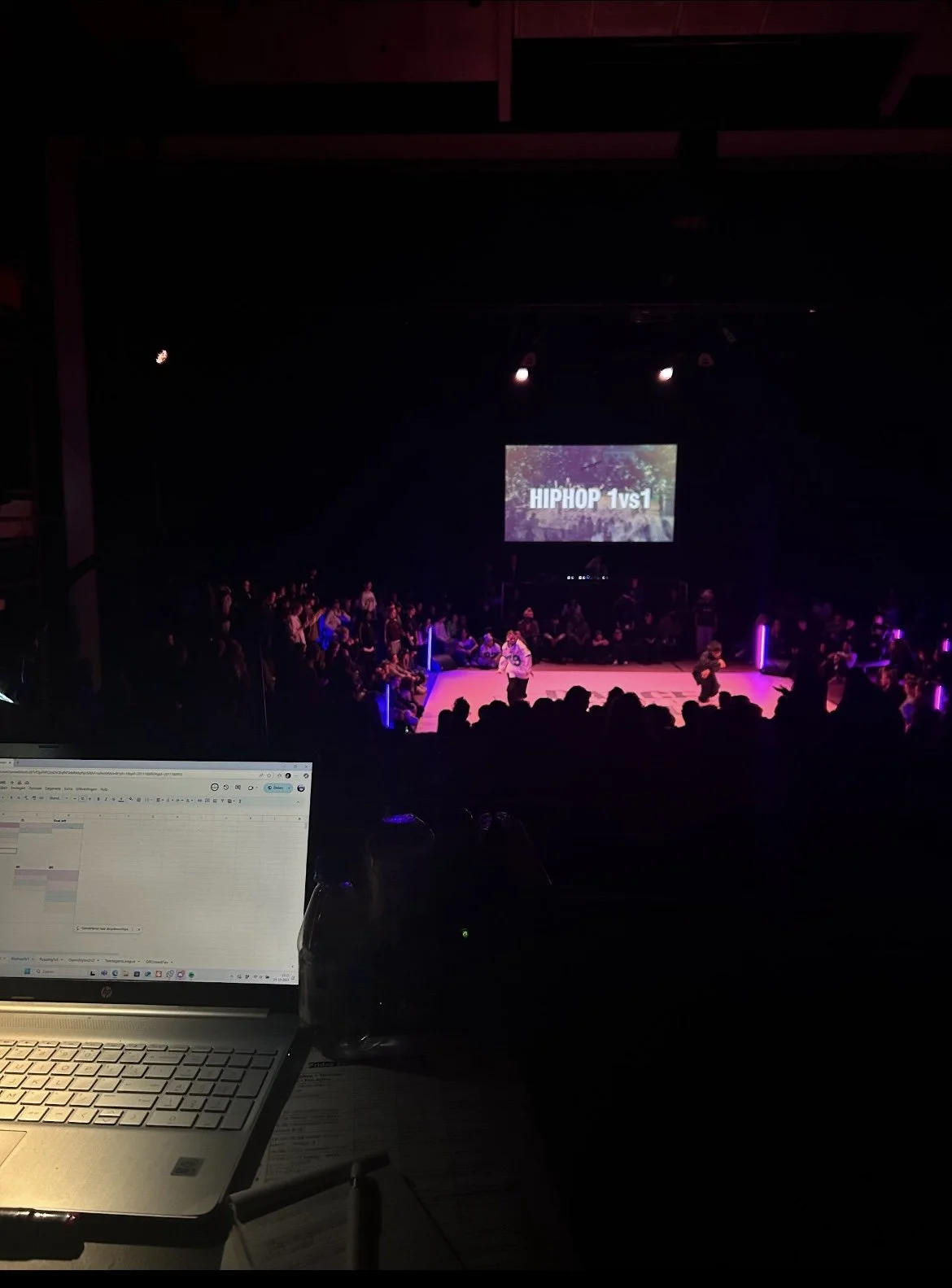 Photo taken from the perspective of a backstage or control area at an indoor hip-hop competition event. In the foreground, there is a laptop on a table, displaying a digital spreadsheet, and a water bottle. The background shows a stage with a large screen displaying 'HIPHOP 1v1', surrounded by an audience seated in a darkened theater. The stage is lit with purple and pink lights, and there are two performers or contestants on the stage.