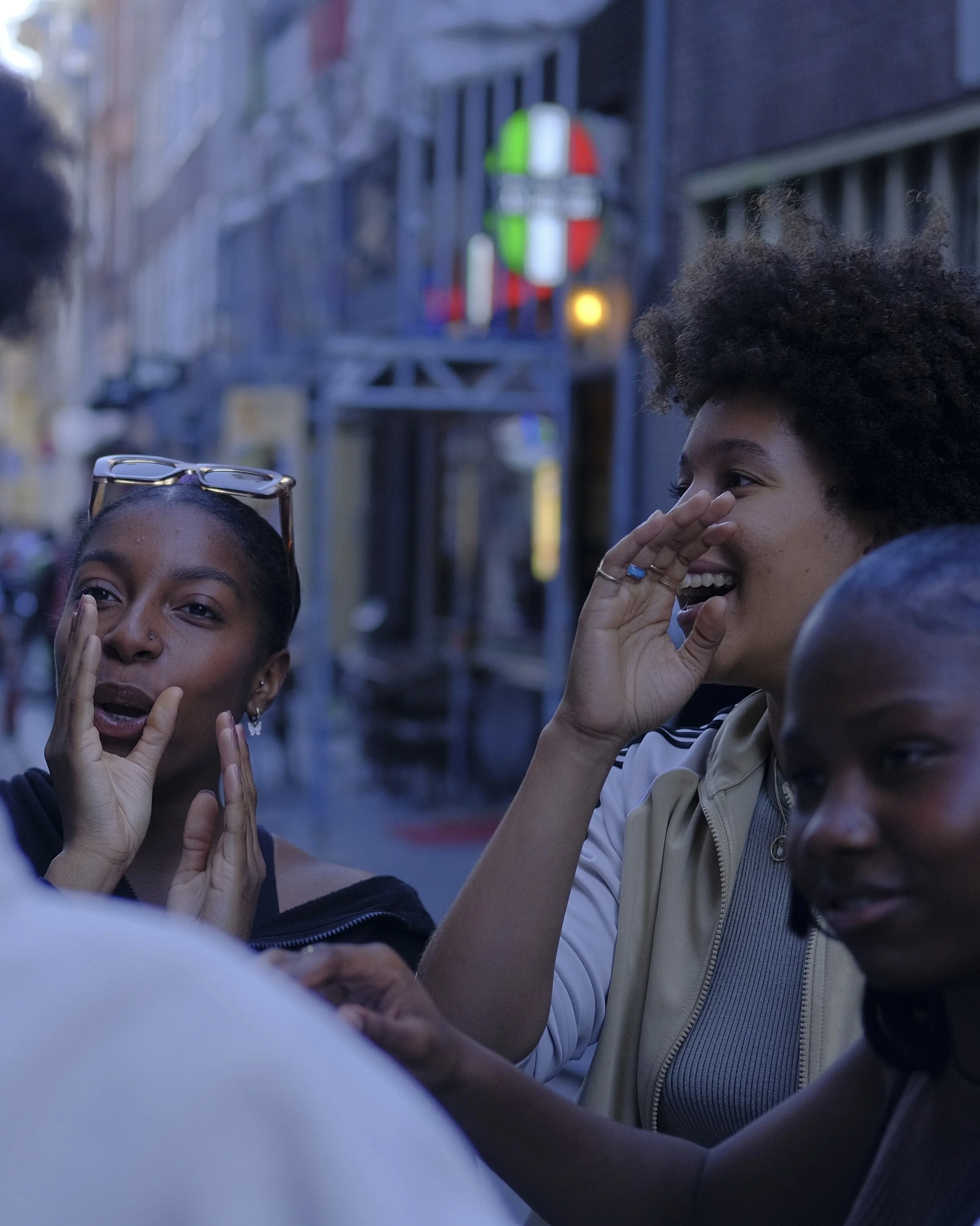 Four women socializing outside in an urban setting during the evening, engaged in conversation and laughter, with a neon sign and buildings in the background.
