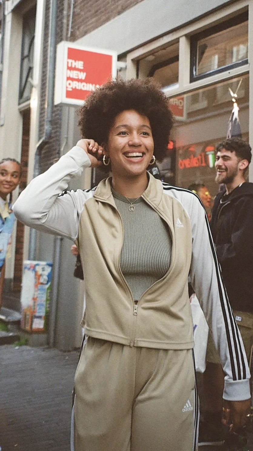 A smiling woman with curly hair wearing a beige Adidas tracksuit, standing on a city street with two people in the background.