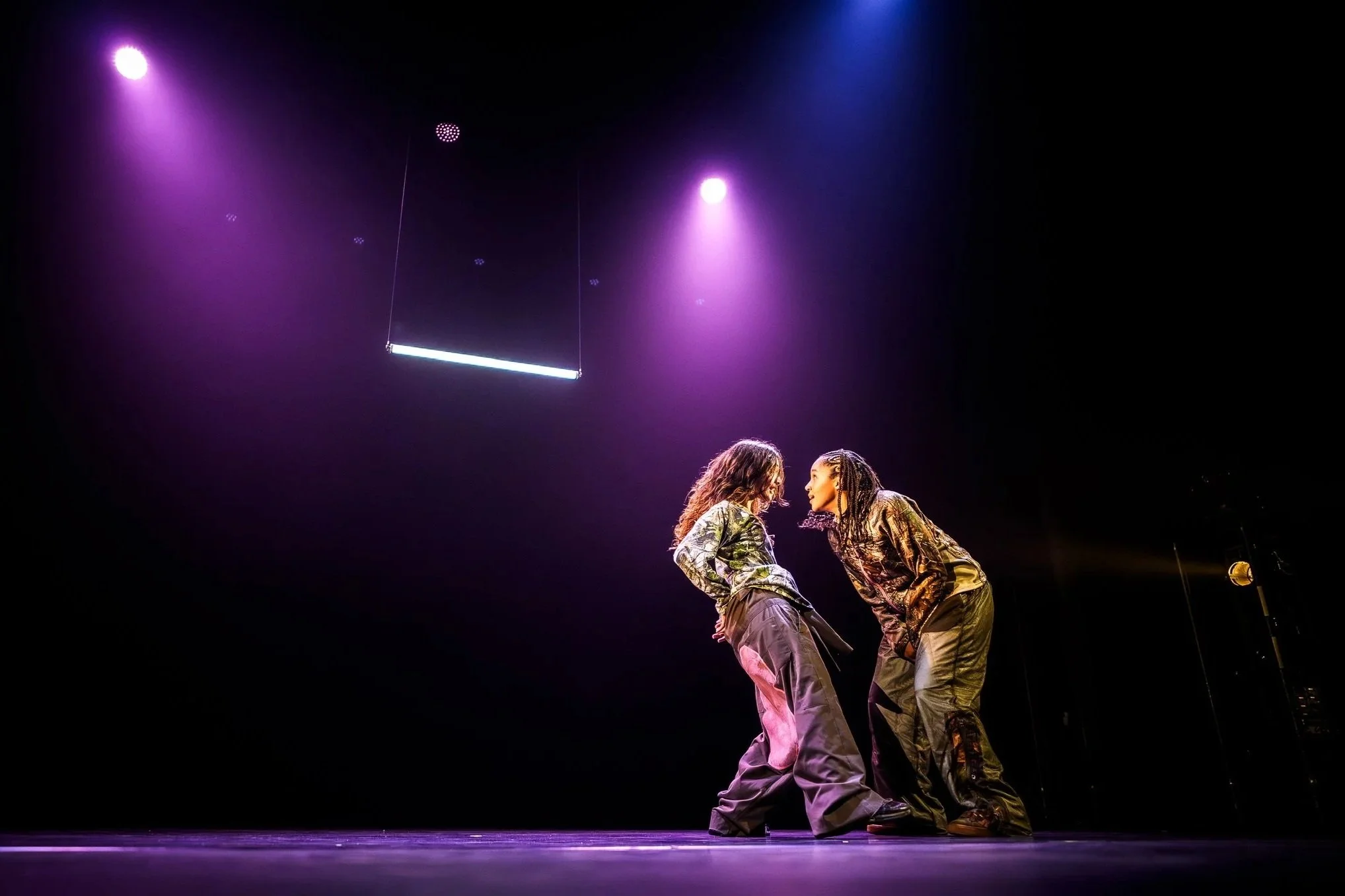 Two performers on stage, leaning towards each other, under purple stage lighting.
