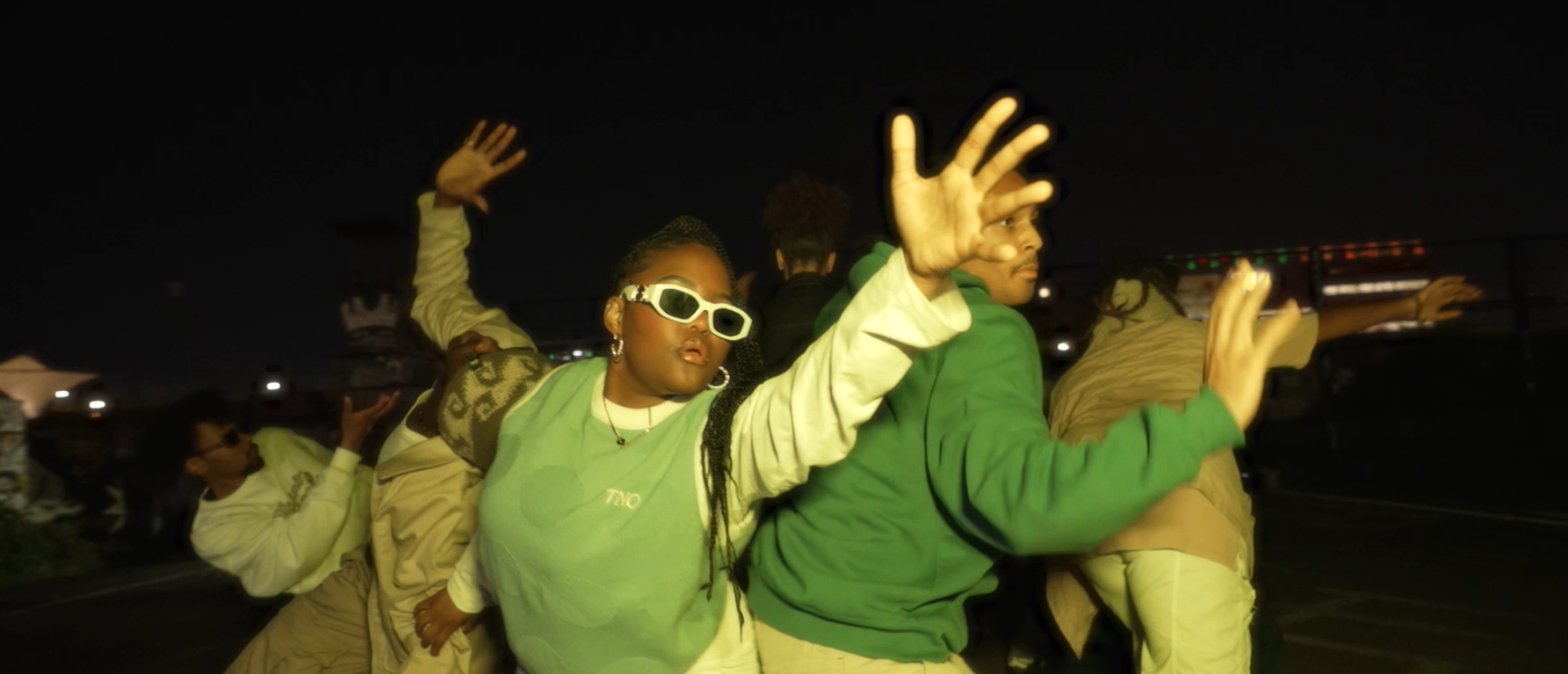 Multiple young people at night, some dancing and posing with hands raised, wearing colorful streetwear and accessories.
