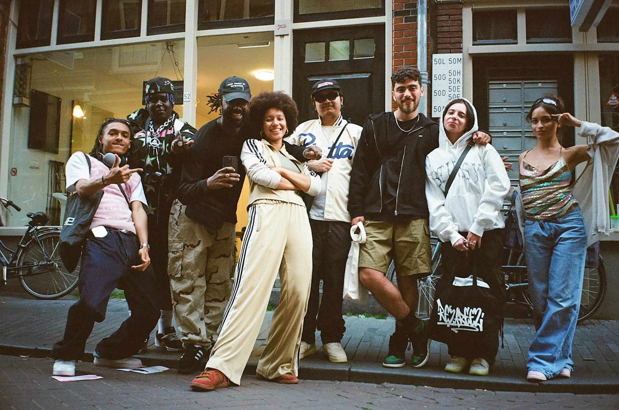 A diverse group of nine young adults standing on a city sidewalk outside a building, posing for a photo. The group includes men and women with various styles, some with casual clothing, others with streetwear. They appear happy and relaxed, with some making gestures or holding items.