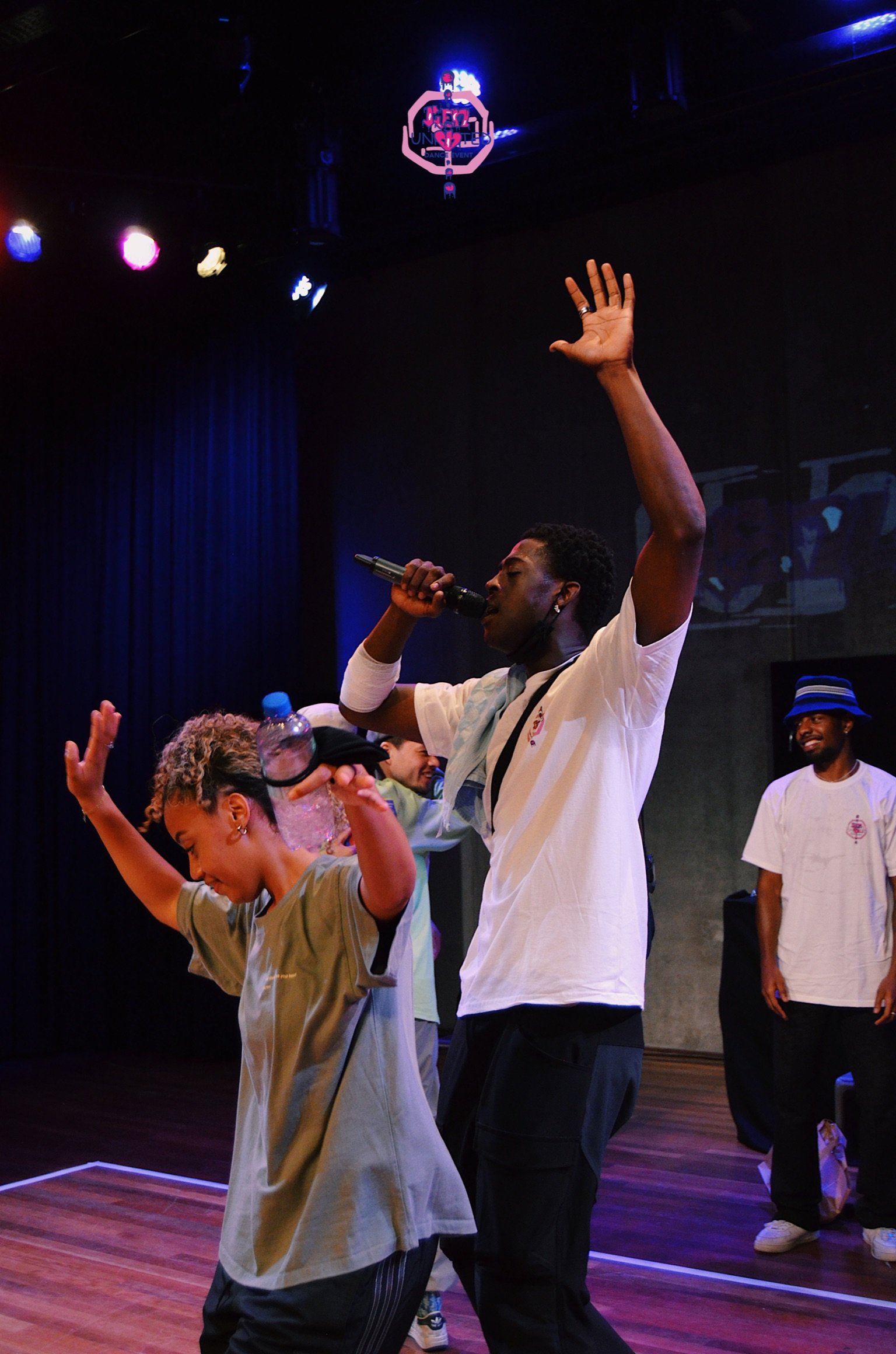 A performer passionately singing on stage with one arm raised, holding a microphone in his other hand, and surrounded by people dancing and enjoying the music.