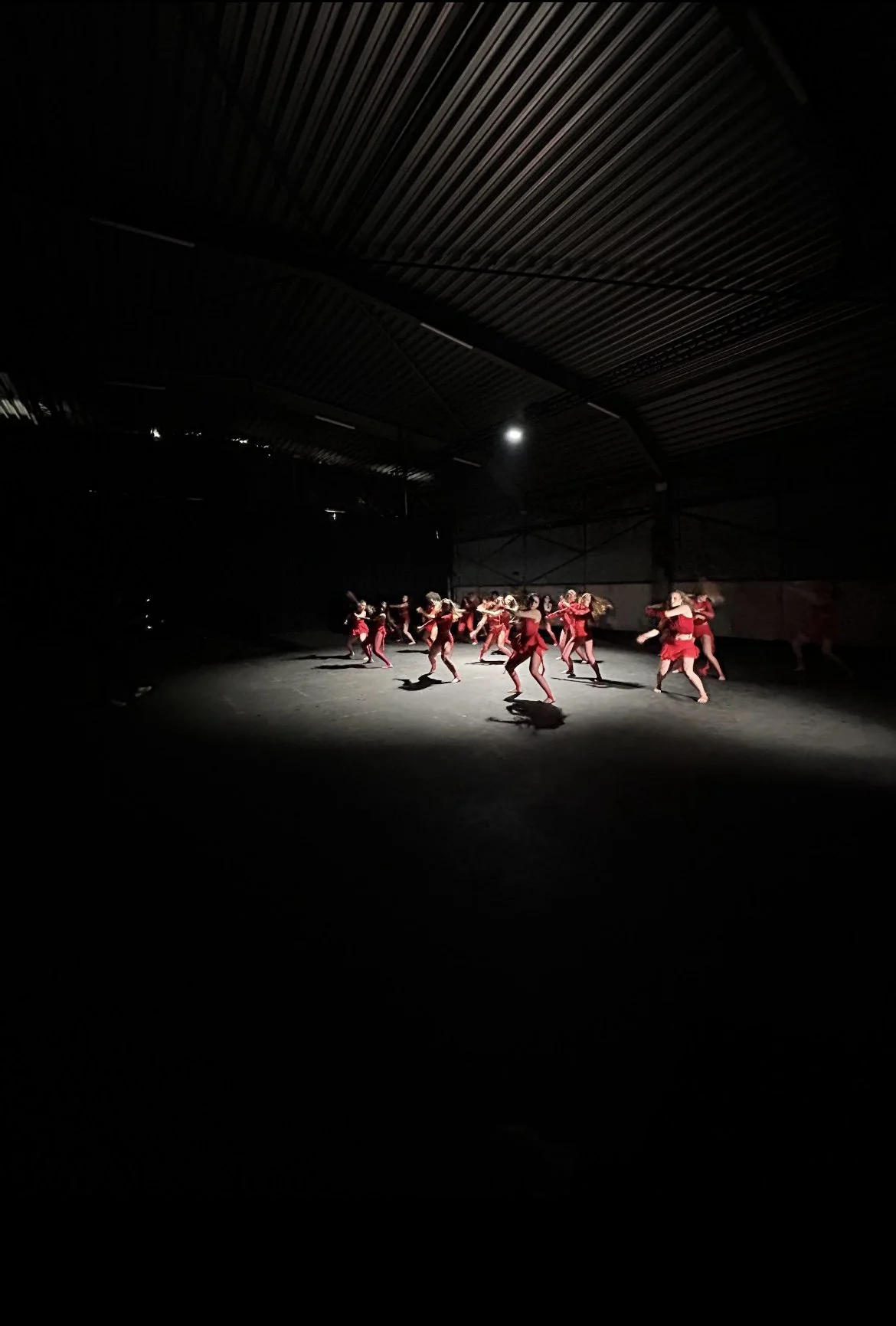 A group of dancers dressed in red performing under a spotlight in a dark industrial space with metal ceiling panels.