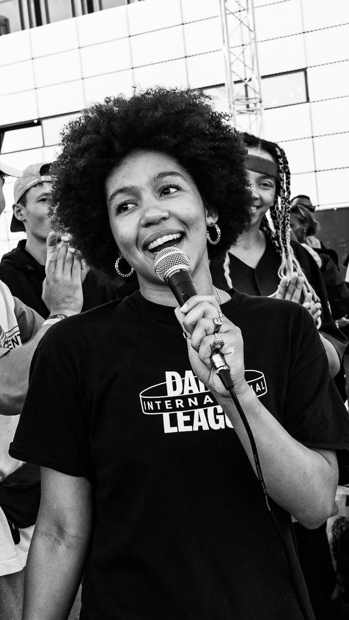 A woman with curly hair and hoop earrings holding a microphone, smiling at an outdoor event with people in the background.