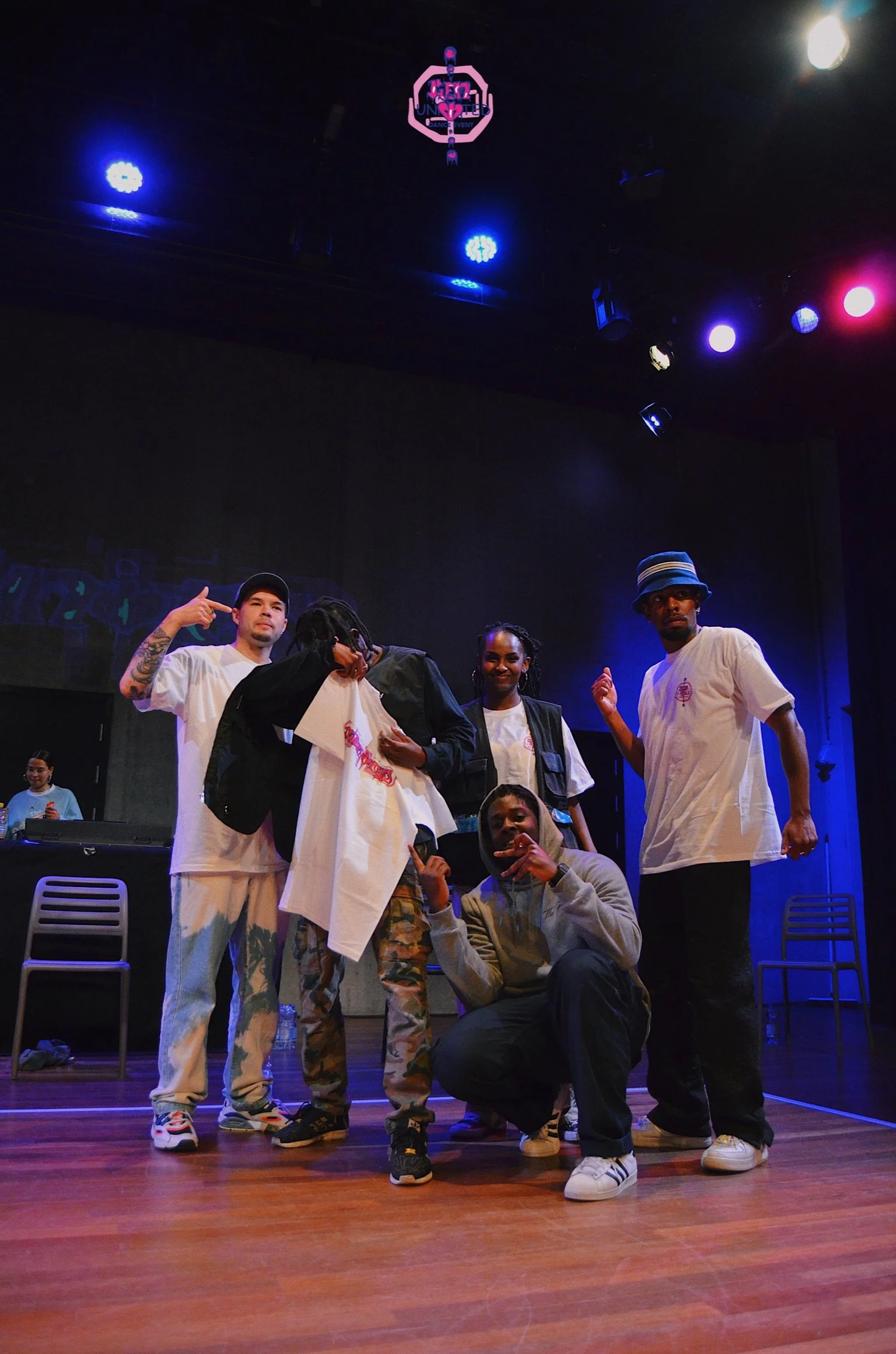 A group of five people on a stage under colorful lights, with one person holding a white T-shirt and other members striking poses. A DJ is visible in the background.