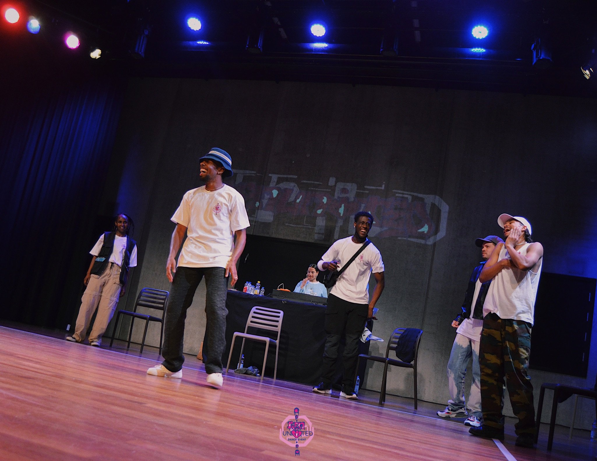 Group of five young people standing and laughing on a stage with a DJ table and chairs, in a dark room with stage lighting.