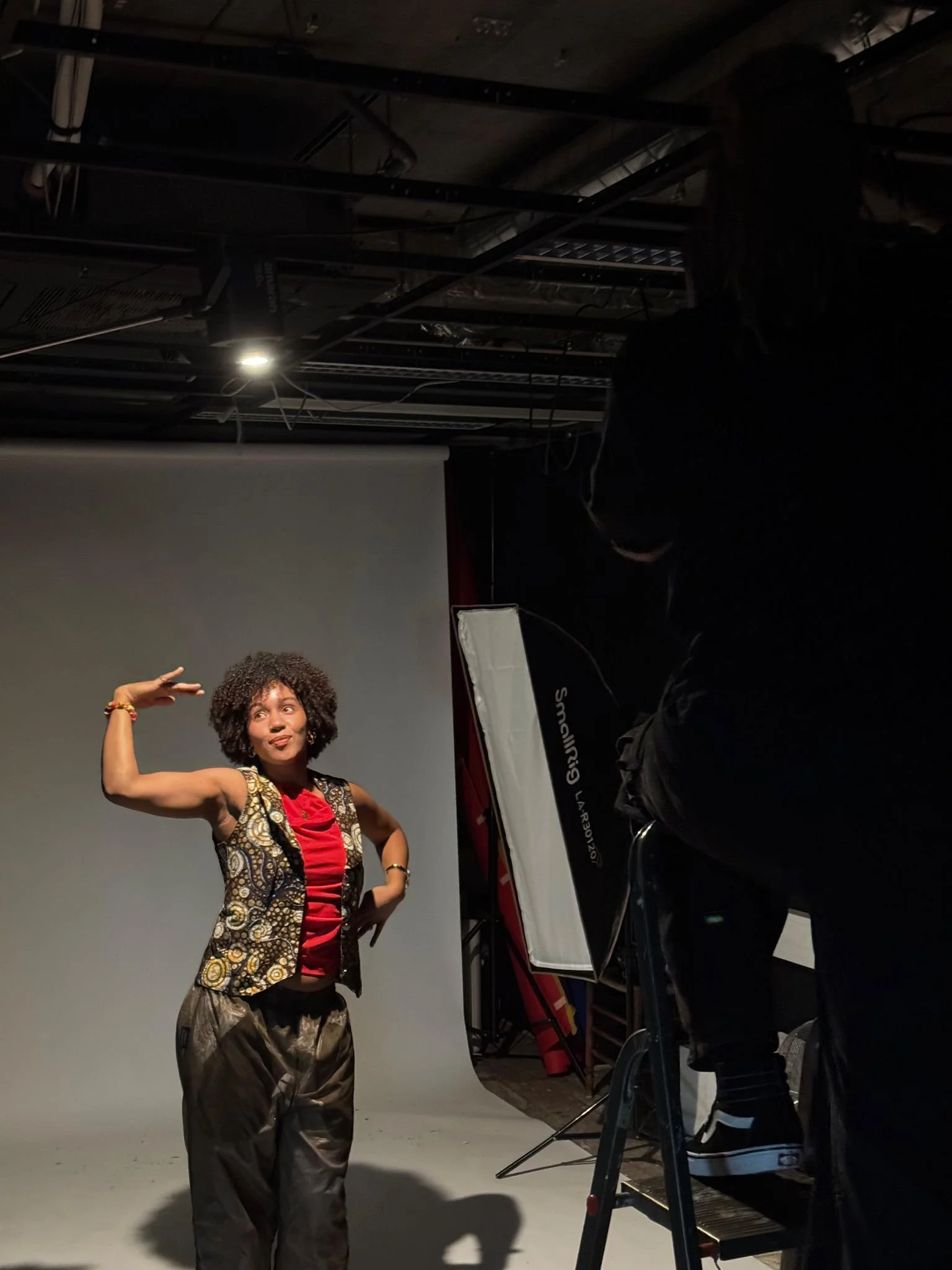 A woman posing for a photo in a photography studio with a gray backdrop, wearing a colorful vest and red shirt, making a gesture with her arm, while a person adjusts equipment nearby.