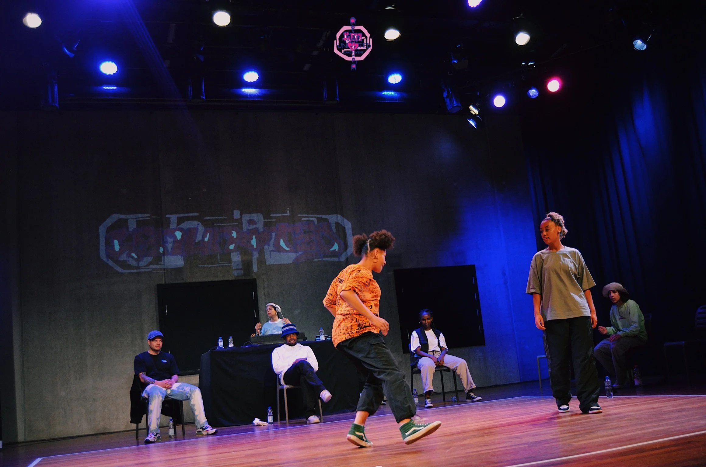Youth performers on stage in a dance or drama rehearsal, with a DJ and audience members seated in the background, under stage lights and dark curtains.