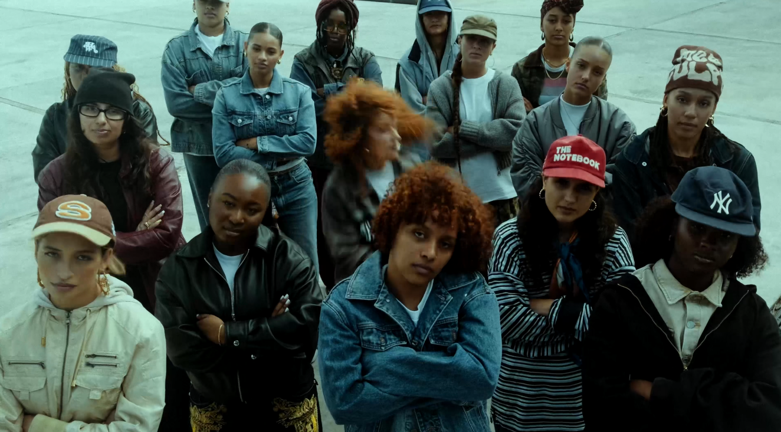 Group of young women standing outdoors with serious expressions, some crossing their arms, dressed in casual streetwear and jackets.