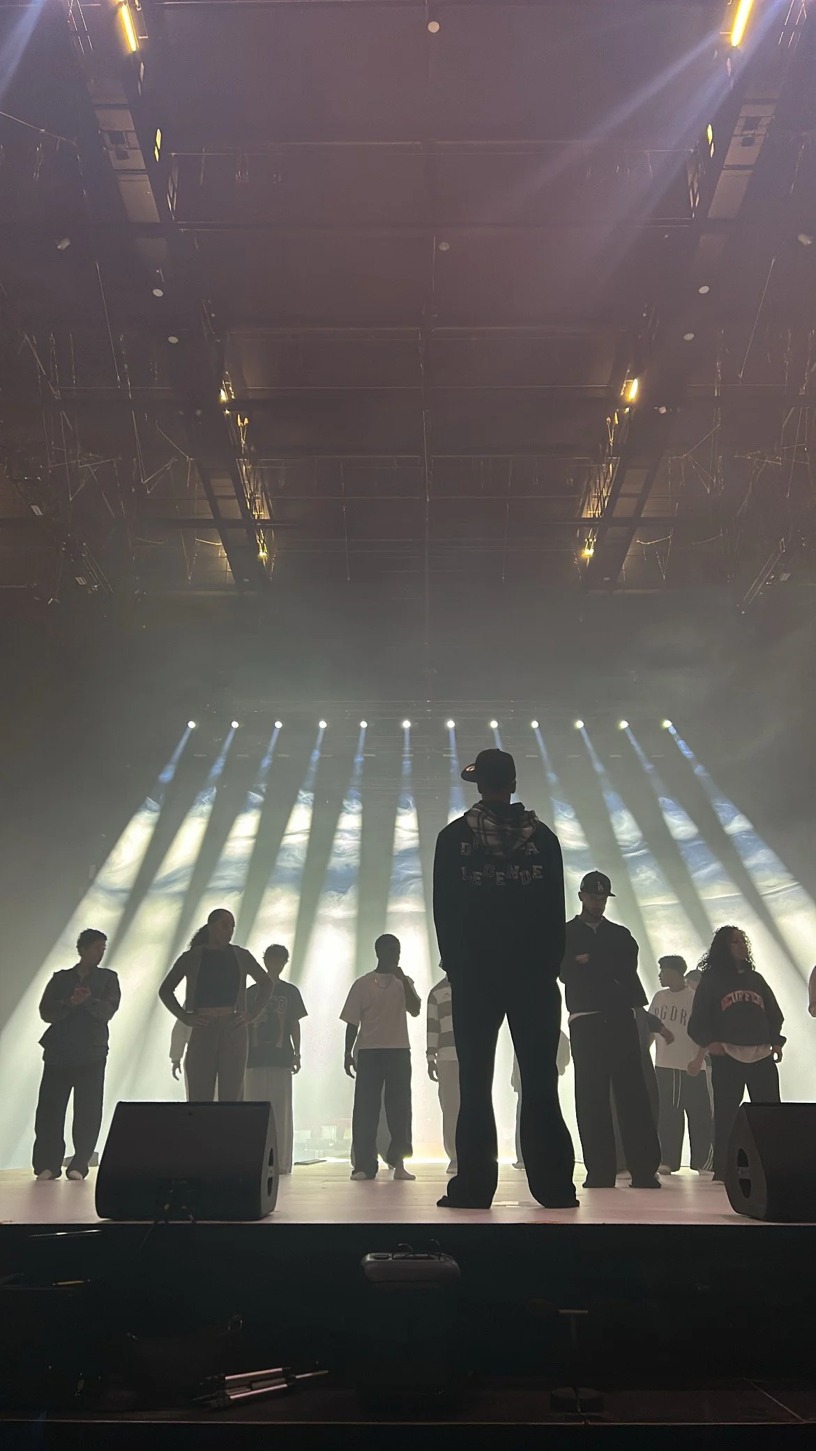 People standing on stage during a rehearsal or sound check with stage lights and smoke effects in the background.