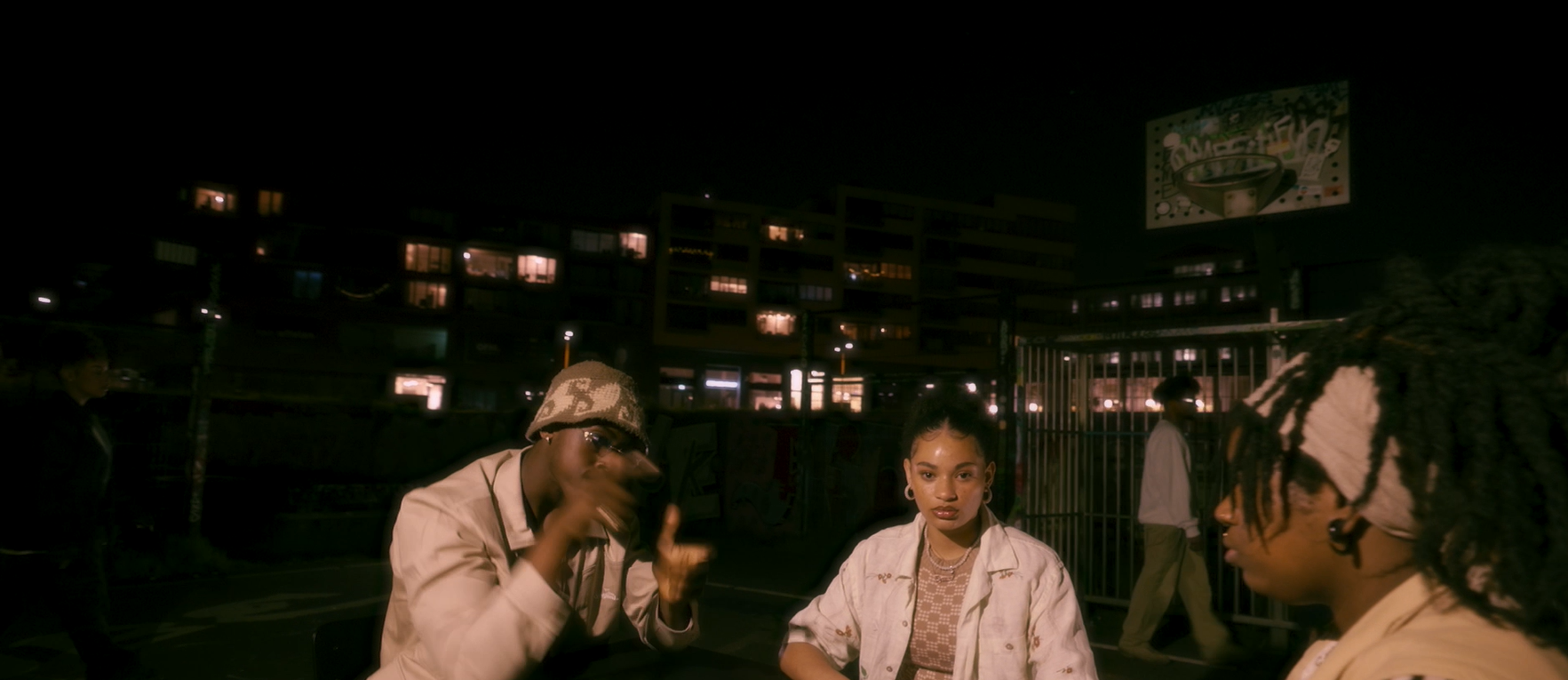 Three young adults sitting outdoors at night, with city buildings and a basketball hoop in the background.