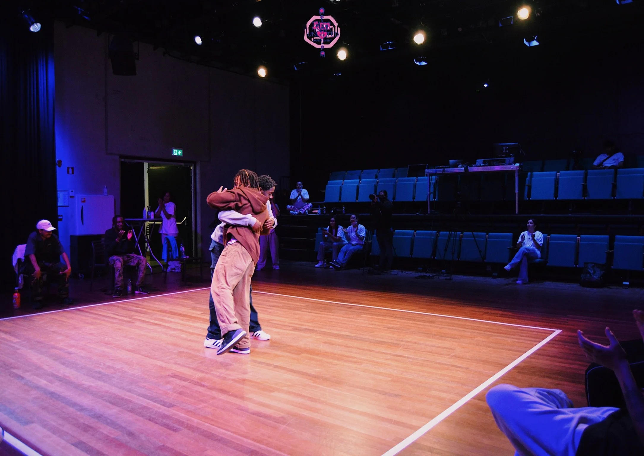 People embracing in the center of a dance studio with a wooden floor, surrounded by seated onlookers, in a dark room with blue lighting and seating.
