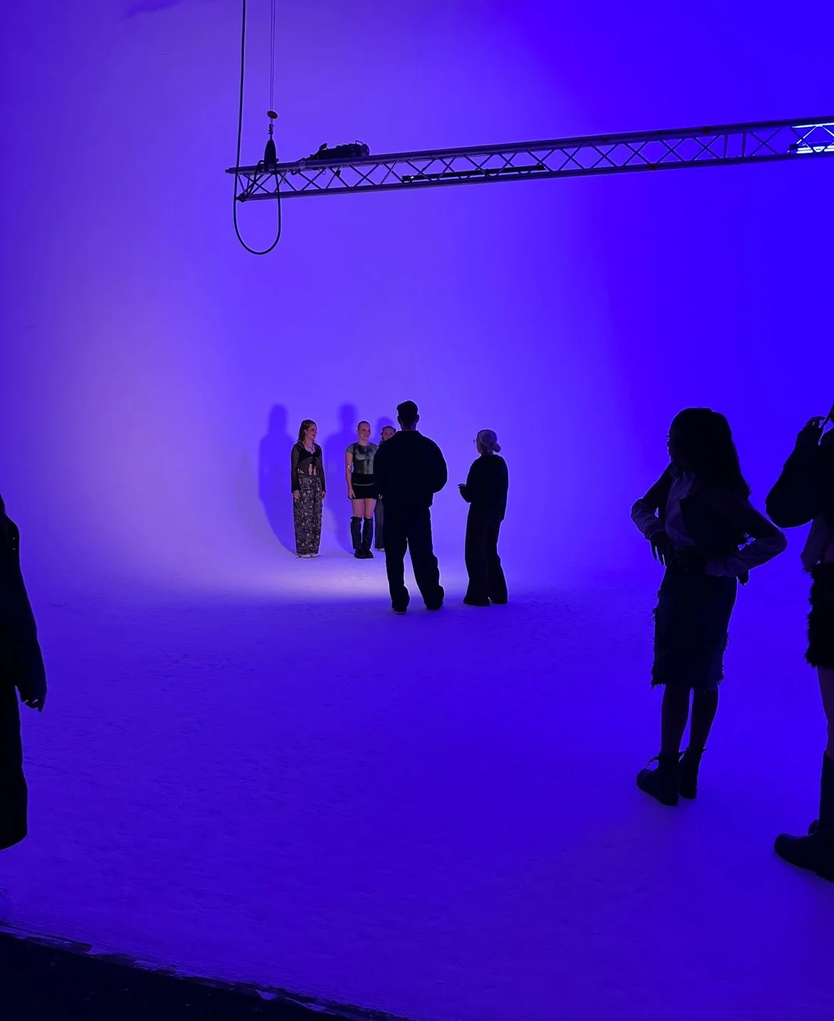 People on a film or photography set with a purple backdrop, some standing and talking, with a lighting rig above.