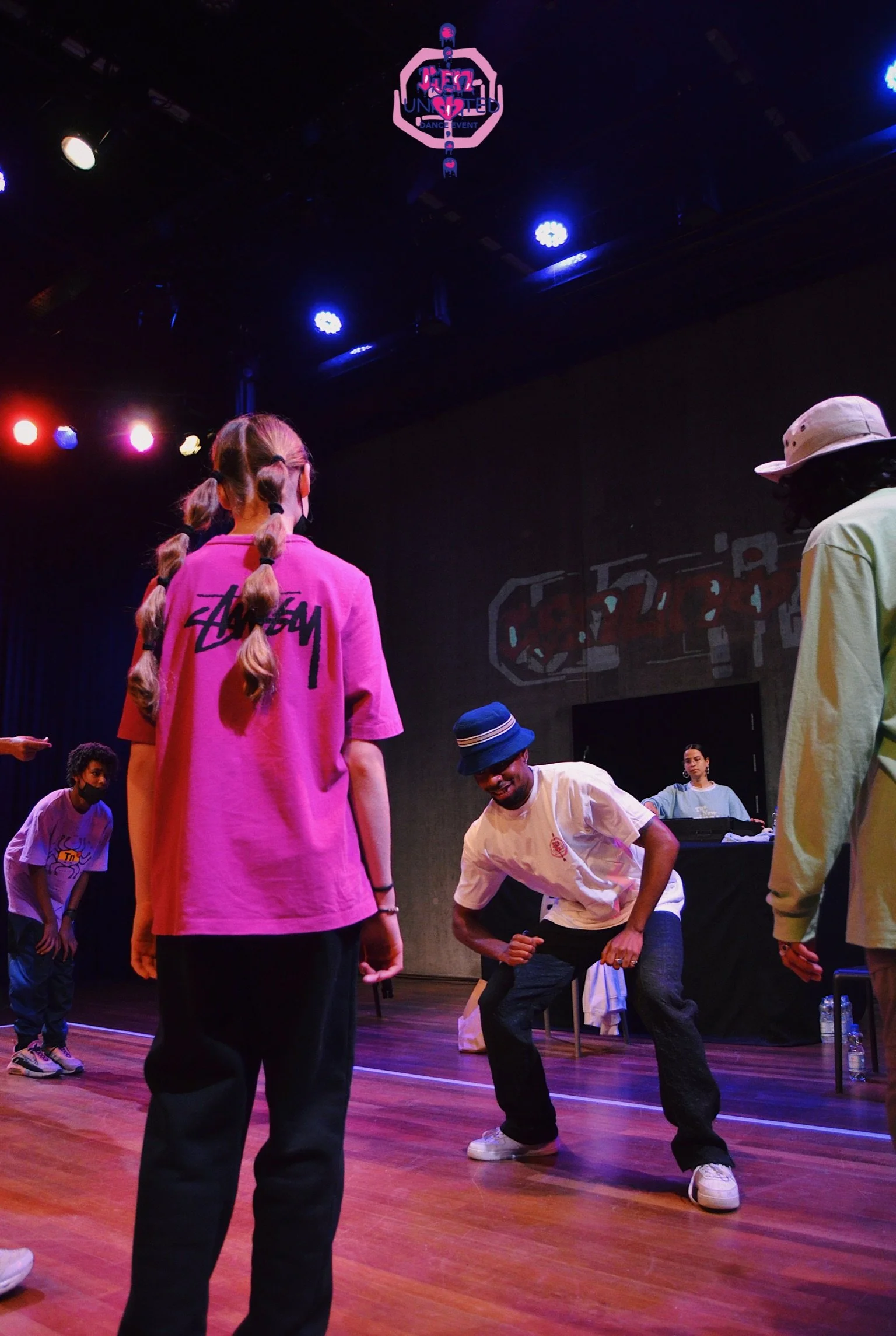 Group of young people dancing on stage at a dance event with stage lights and a DJ in the background.