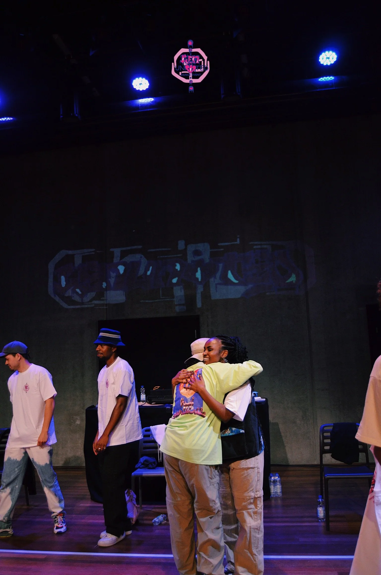 Two people hugging on stage, surrounded by a group of people standing in a line. Dark background with stage lights, DJ equipment, and water bottles on the floor.