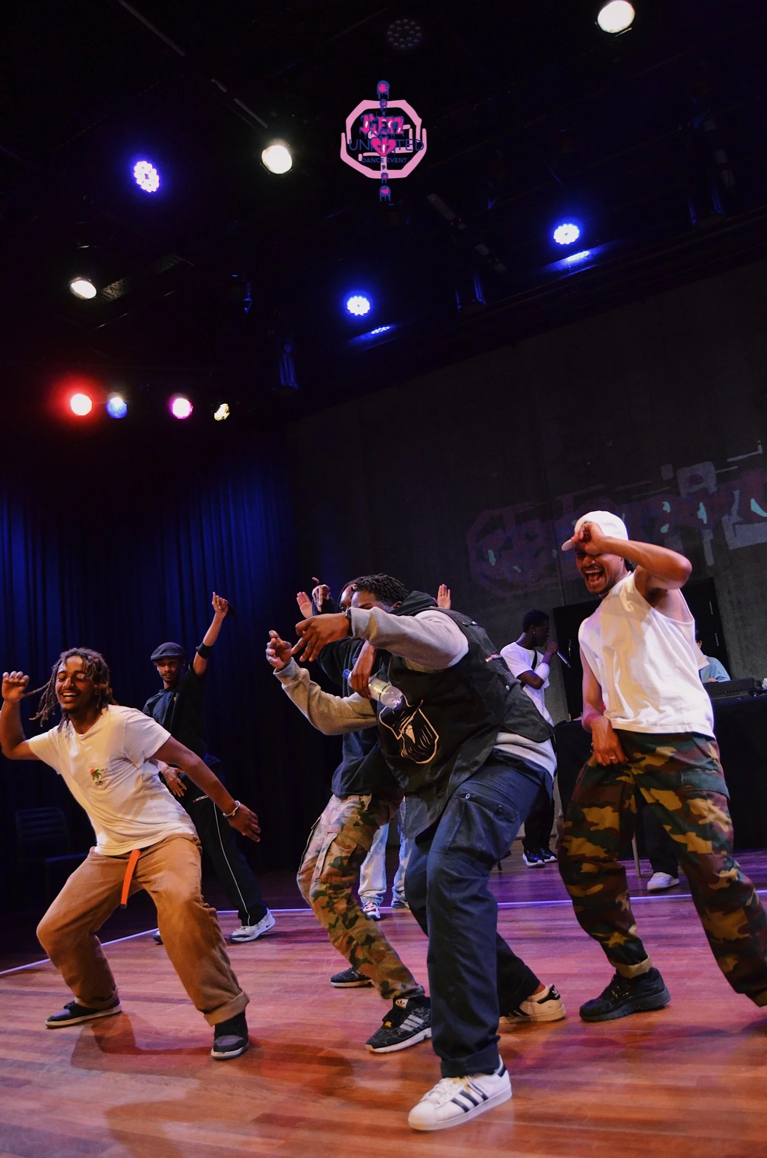 Group of people dancing on stage with colorful lighting, some wearing casual clothes, in a lively dance event.