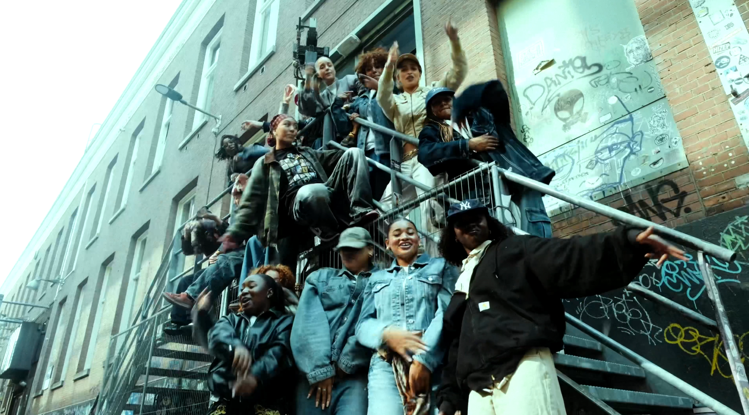 A diverse group of young people gathered on outdoor metal stairs in an urban setting with graffiti on the wall behind them, some waving and smiling.