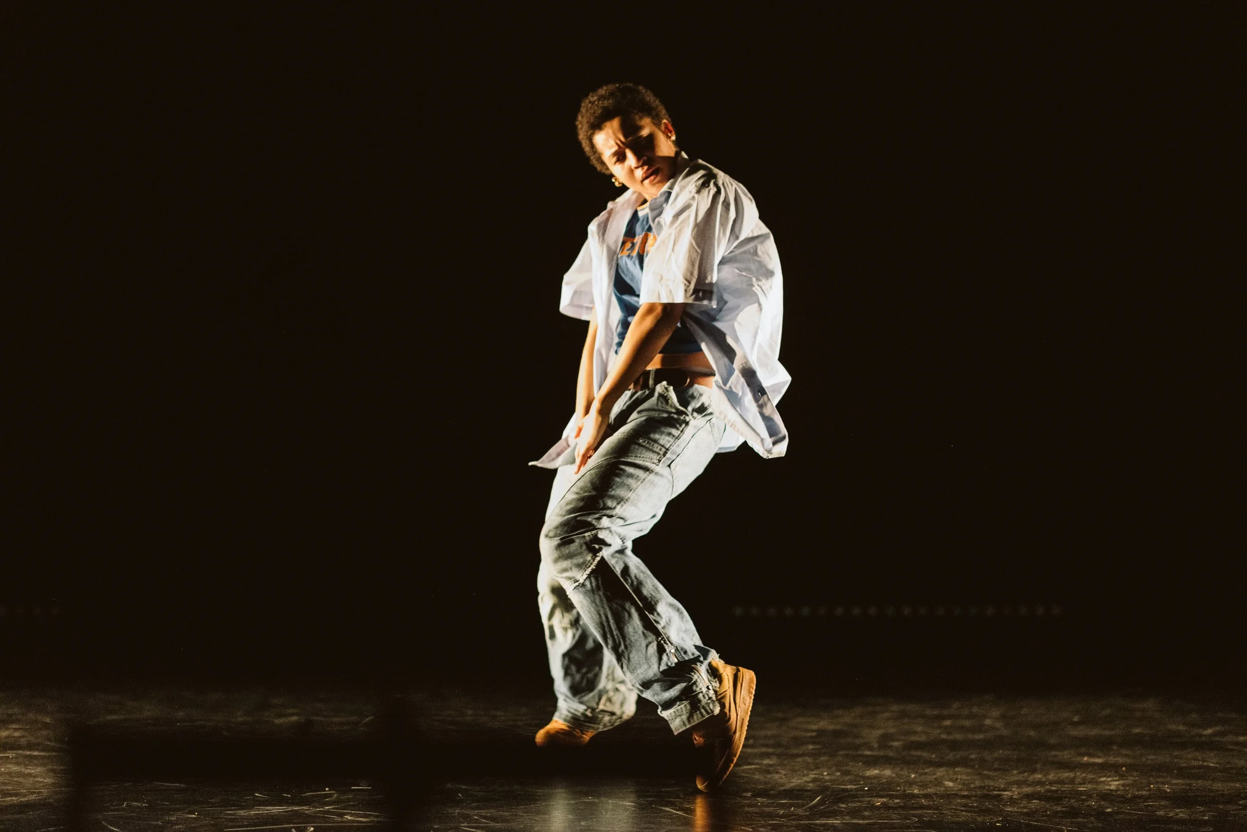 Young person dancing on a dark stage, mid-motion, wearing a white oversized shirt, jeans, and brown shoes, with a concentrated expression.