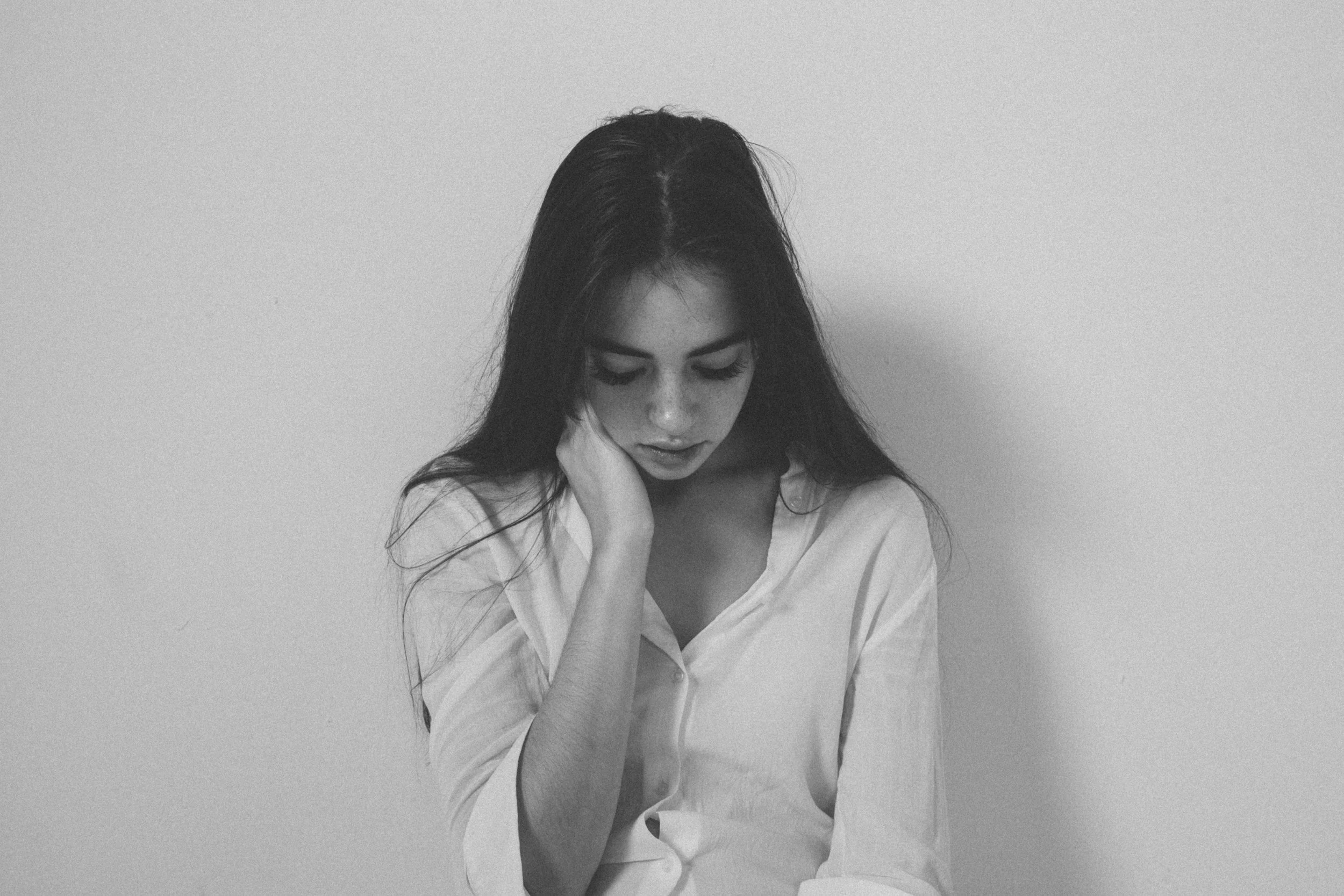 Black and white photograph of a woman with long dark hair, wearing a white shirt, looking down with her hand resting on her face against a plain background.