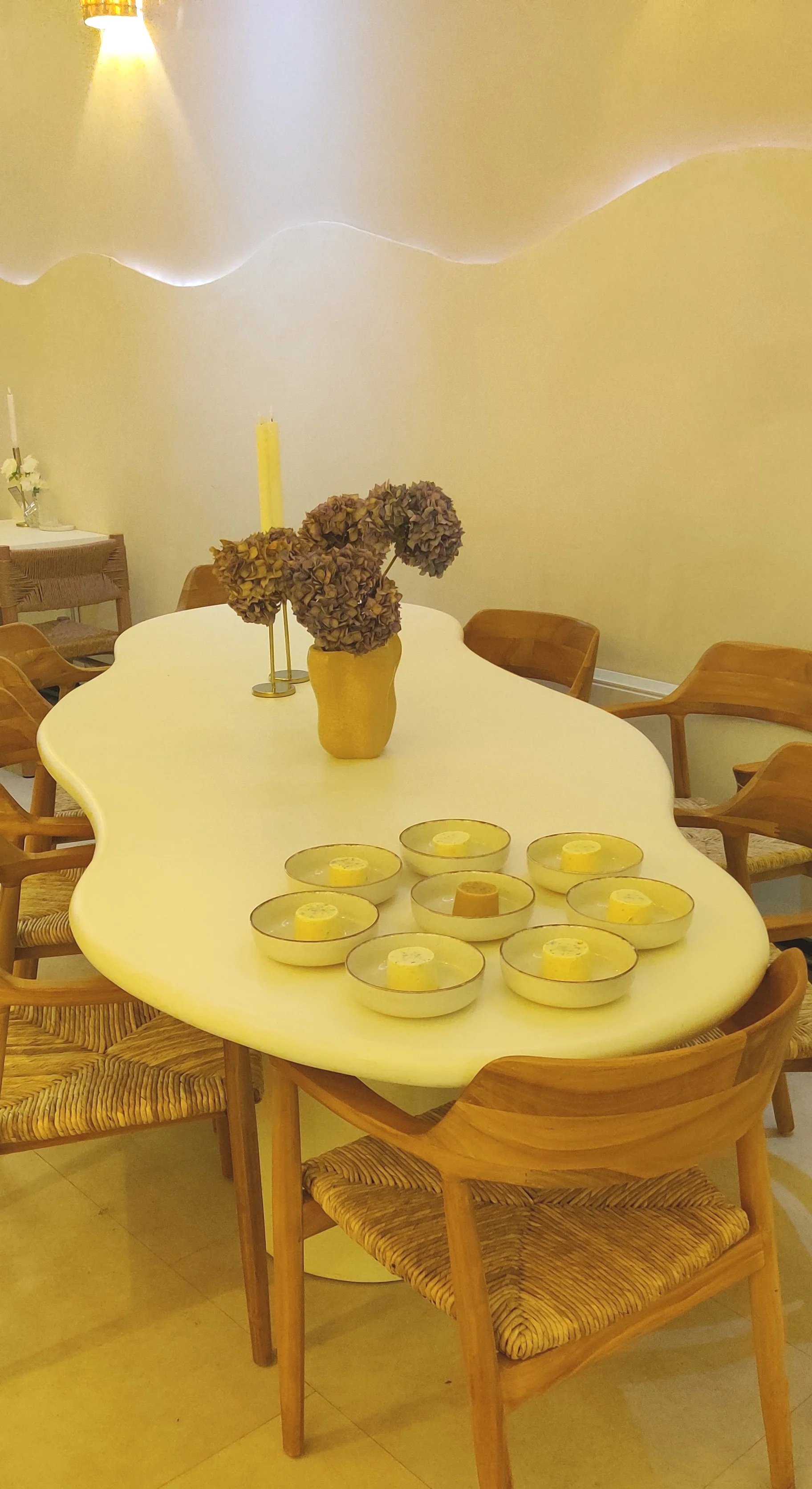 A dining room with a large white, wavy-edged table surrounded by wooden chairs with woven seats. On the table, there are nine small yellow candles in glass holders, a vase with dried hydrangea flowers, and two tall yellow candles in candle holders. The wall behind has soft yellow lighting, creating a cozy atmosphere.