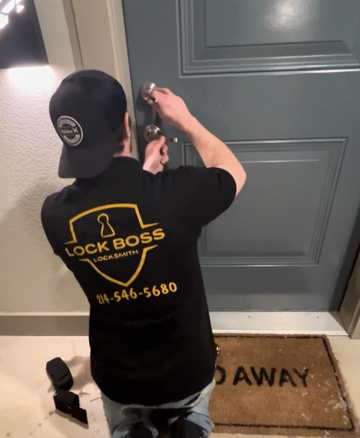 A locksmith kneeling in front of a door, adjusting the doorknob. The person is wearing a black cap and a black T-shirt with yellow print that reads 'LOCK BOSS LOCKSMITH' along with a phone number. There is a mat in front of the door that says 'BACK AWAY'.