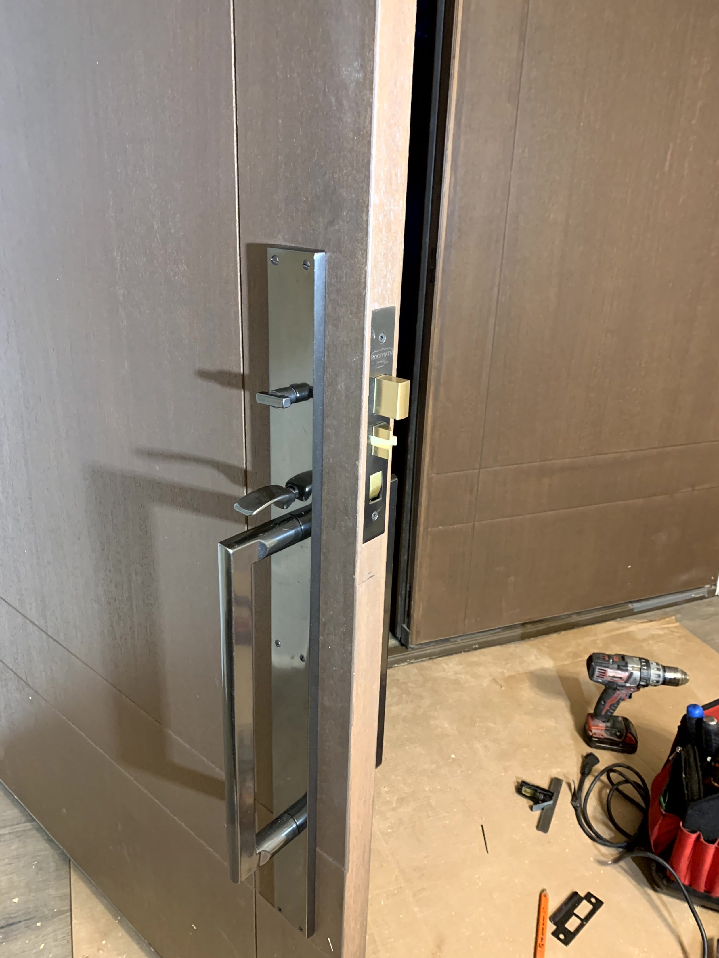 Close-up of an installation of a fire door with panic hardware, a metal door handle, and locking mechanisms, with tools and equipment on the floor nearby.