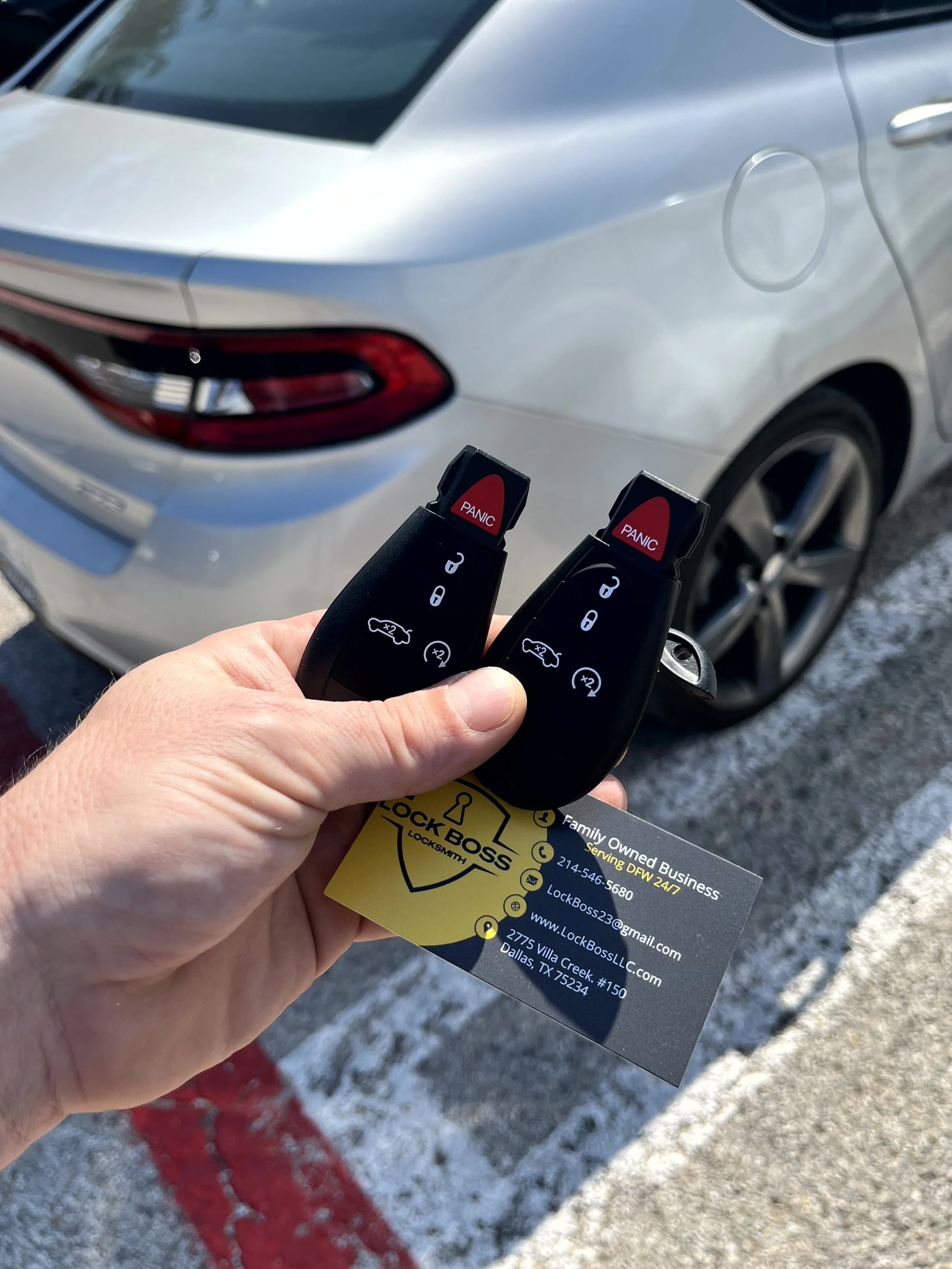 Person holding two car key fobs and business card in front of a silver car parked on a snowy surface.