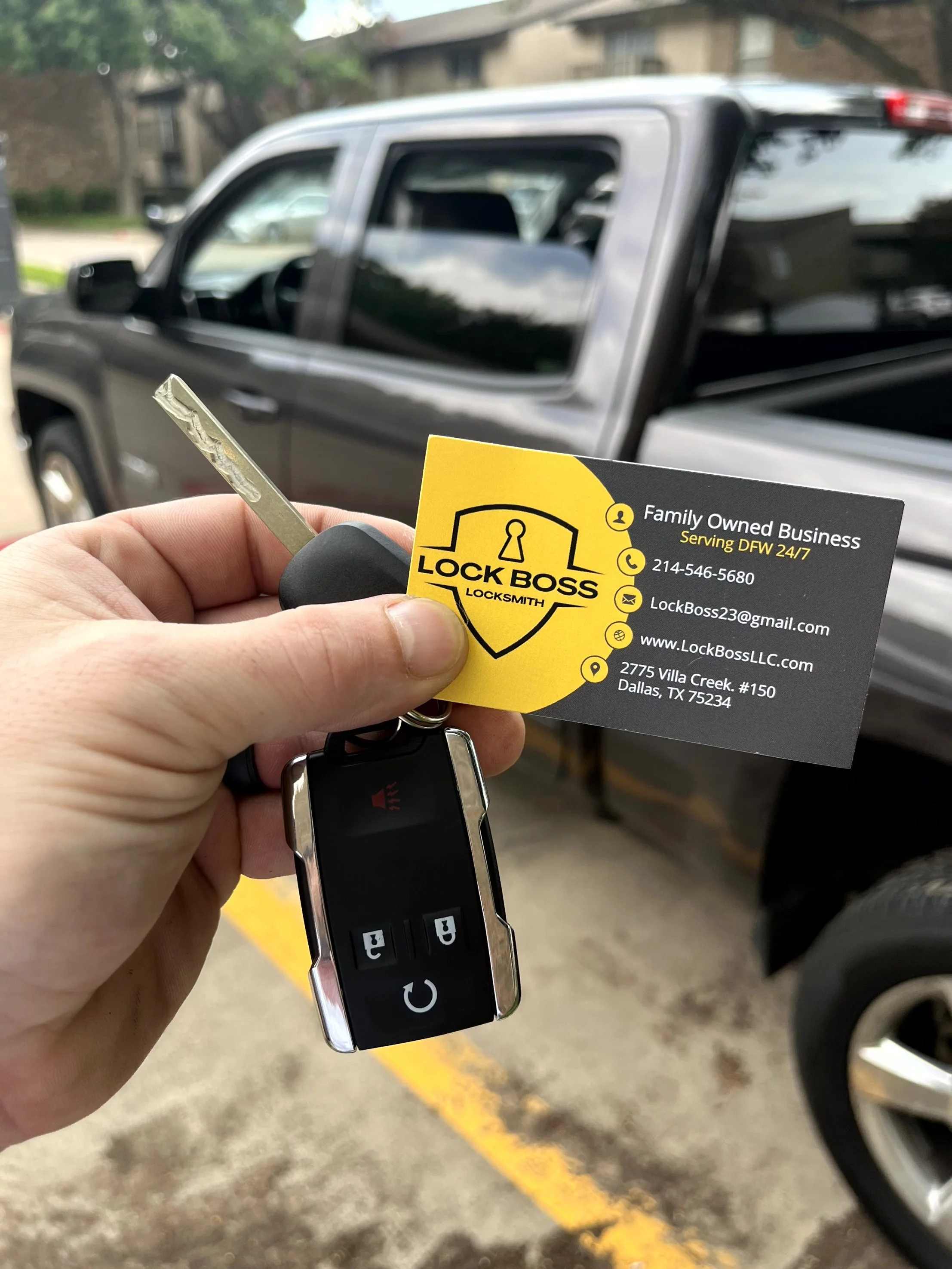 Person holding a car key fob and a business card for Lock Boss locksmith in front of a black pickup truck in a parking lot.