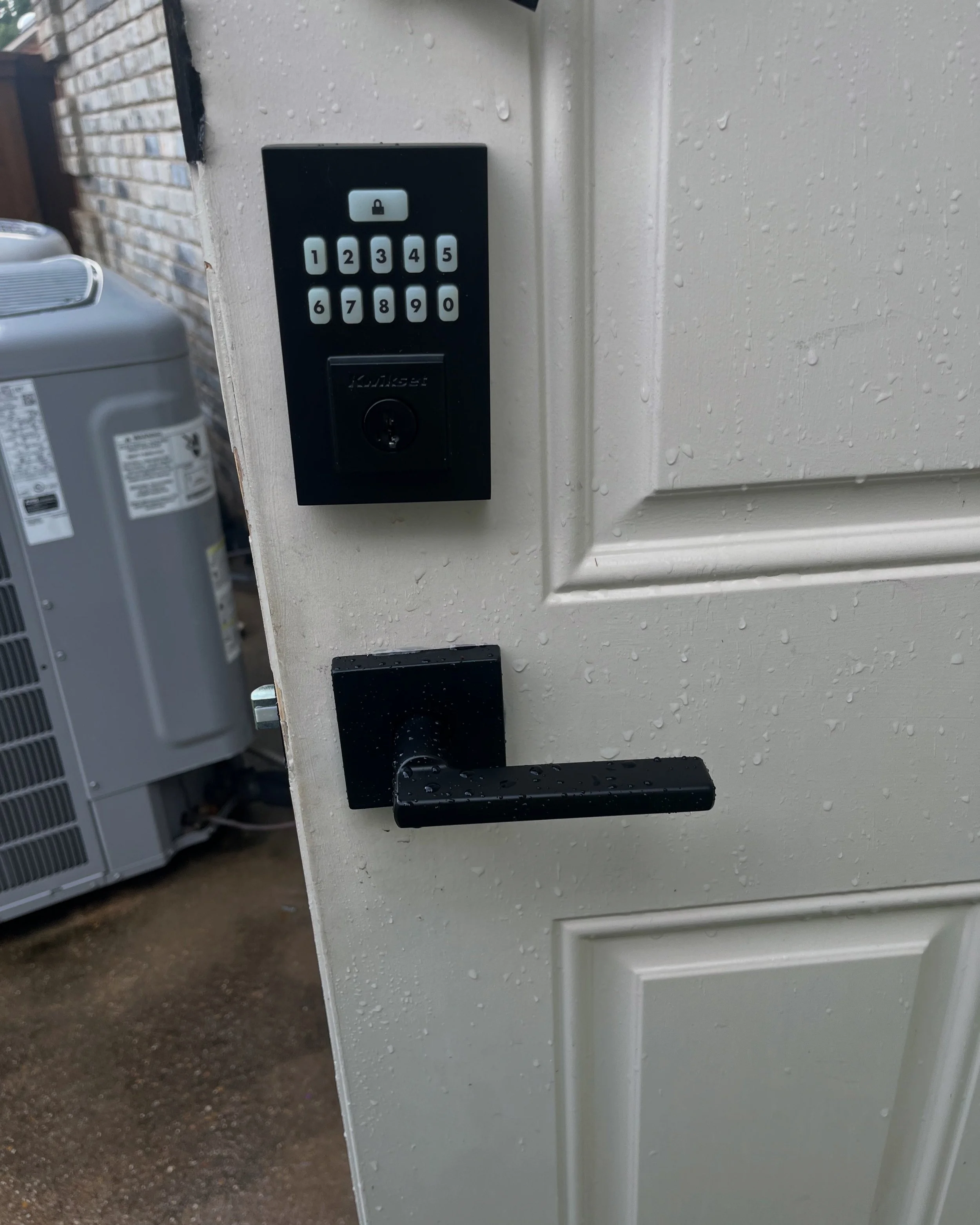 Resistant white door with black electronic keypad lock and handle, and rain droplets on the surface; background shows an air conditioning unit and brick wall.