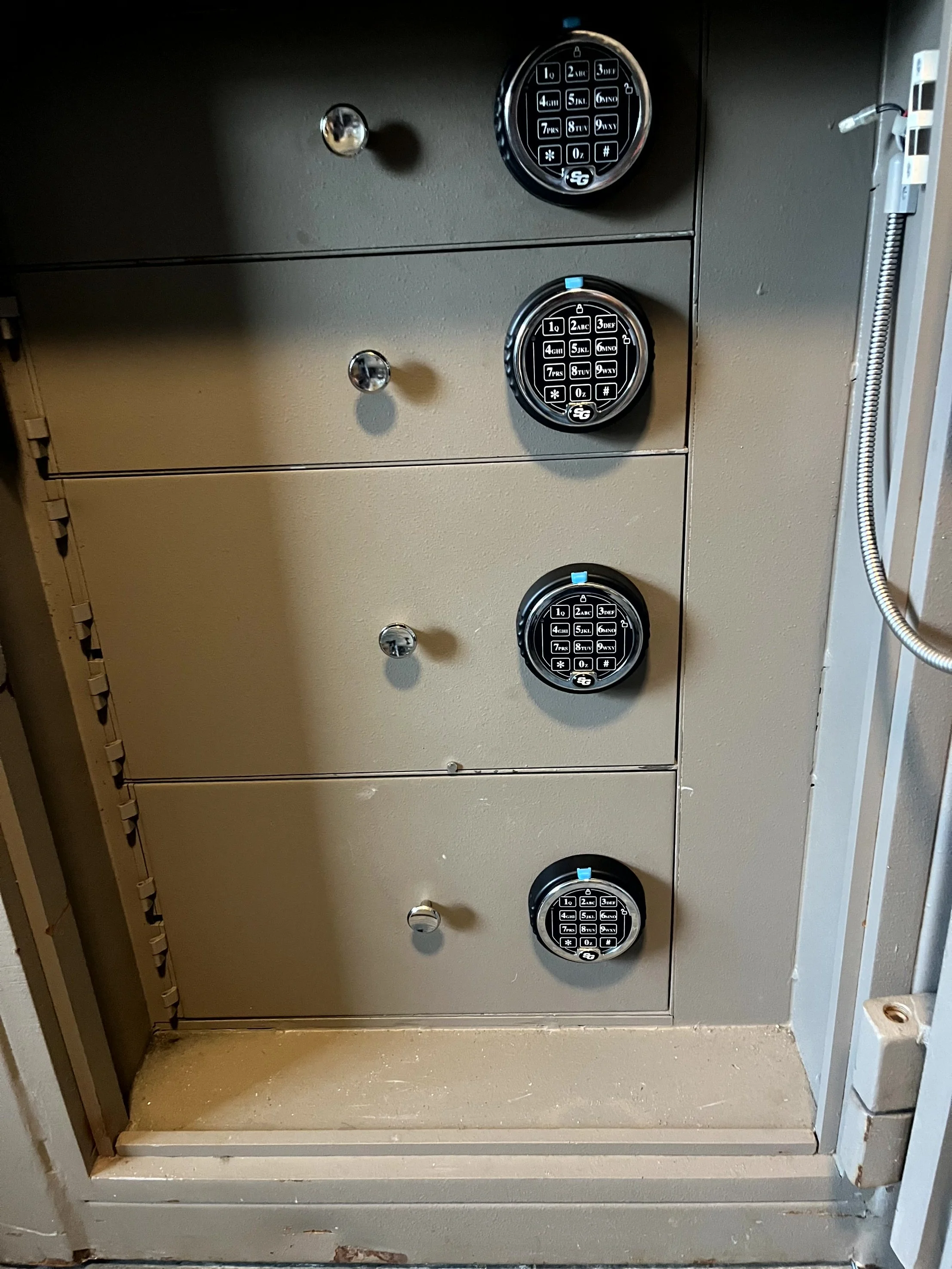 A security safe with four digital keypad locks, three of which are black with blue indicators, inside a beige metal cabinet.