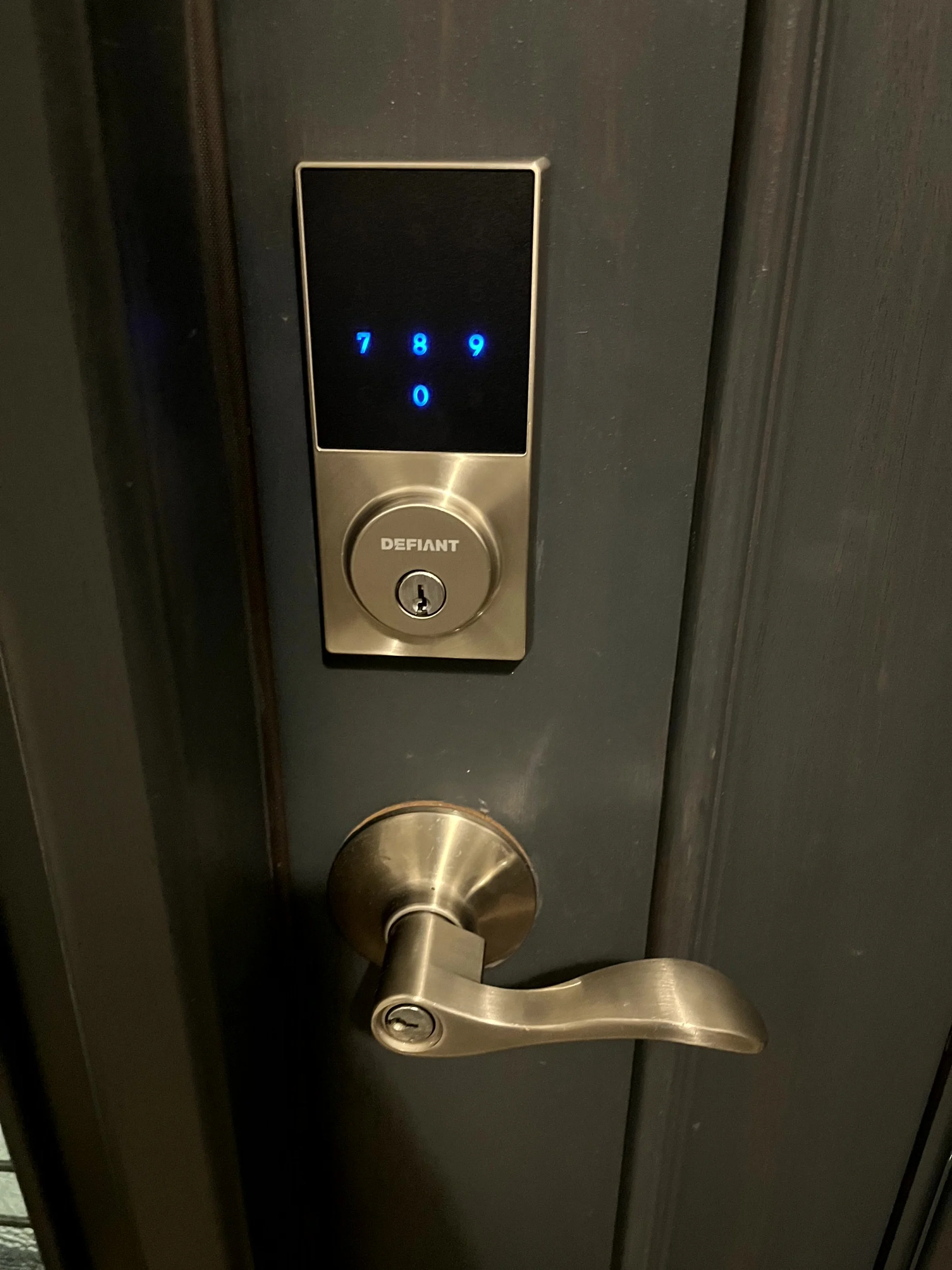 Digital keypad lock with a screen displaying numbers 7, 8, 9, and 0, and a keyhole below it, installed on a dark-colored door with a metallic handle.