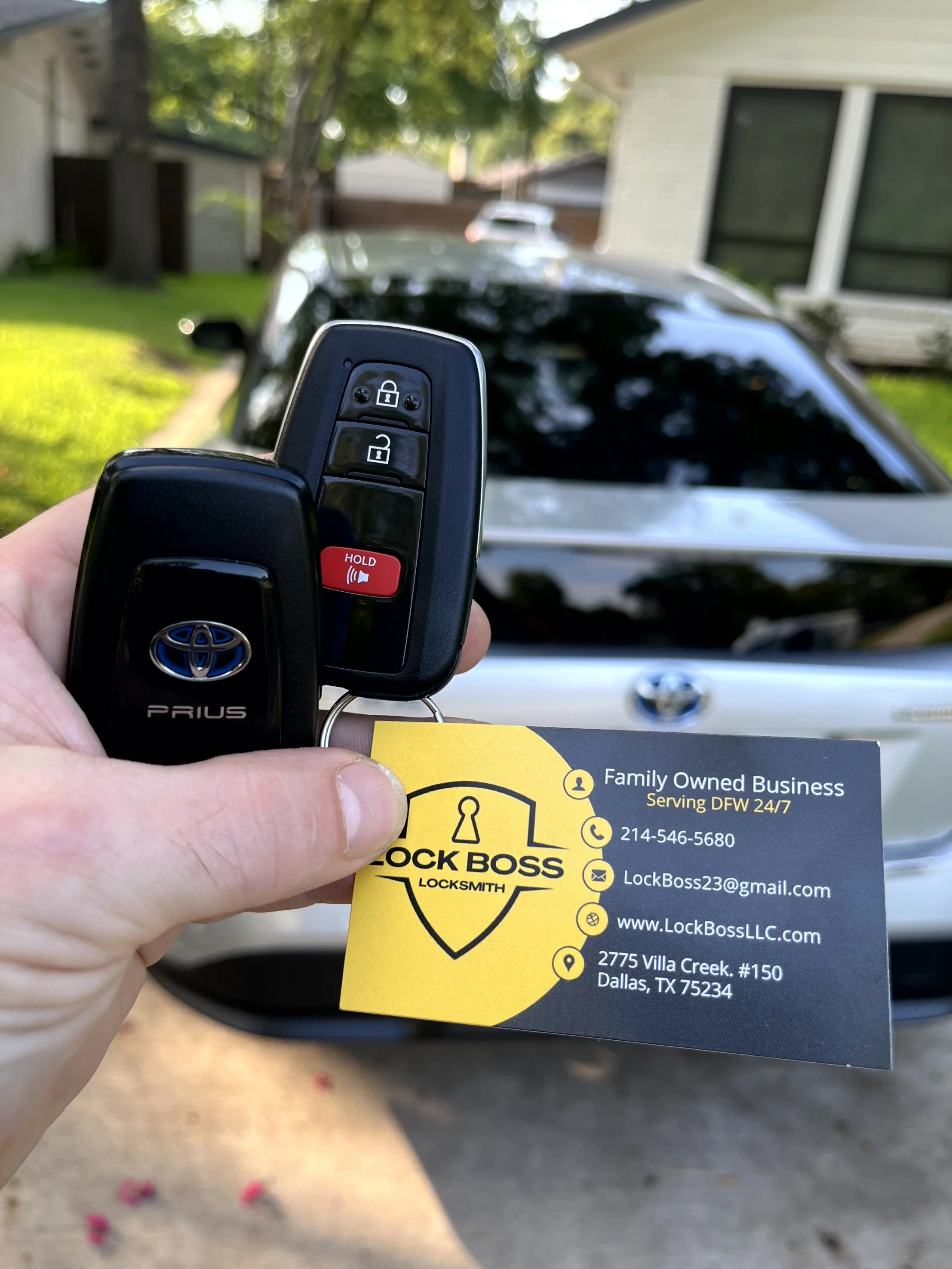 Hand holding Toyota Prius key fob and locksmith business card in front of parked Toyota Prius in a residential driveway.
