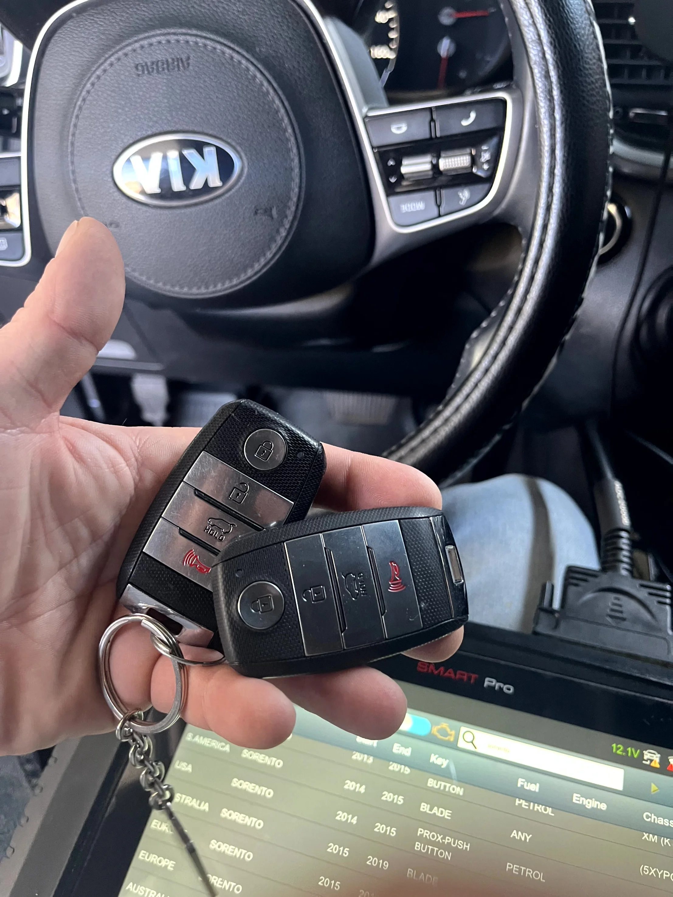 Close-up of a hand holding two car key fobs with a car dashboard and a diagnostic scanner in the background.