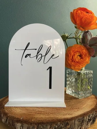 Table number sign with the number 1 and the word table in a script font, next to a glass vase with orange ranunculus flowers on a wooden surface.